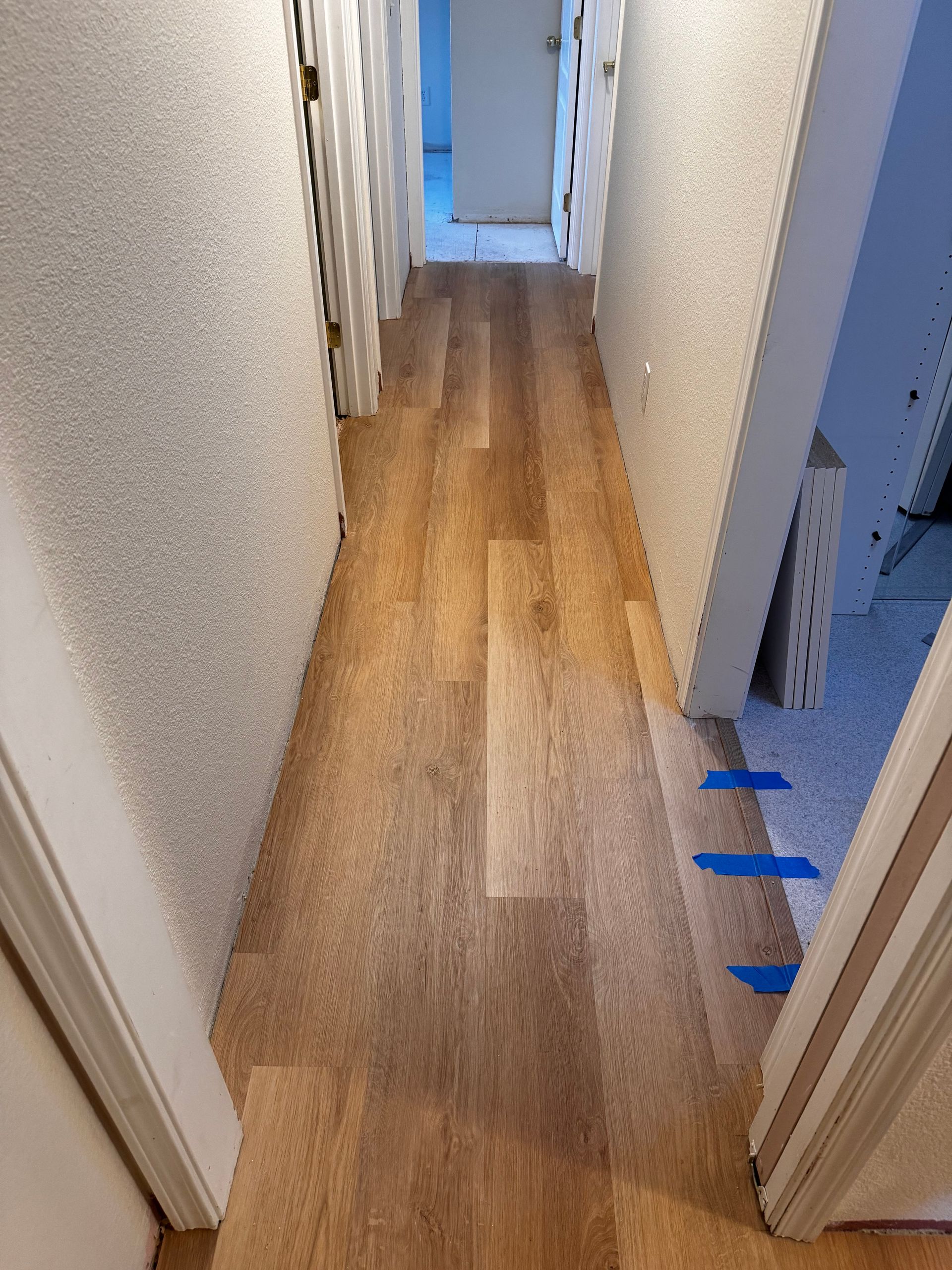 Long hallway with wood-look flooring. White textured walls and doorways. Blue tape marks the floor.