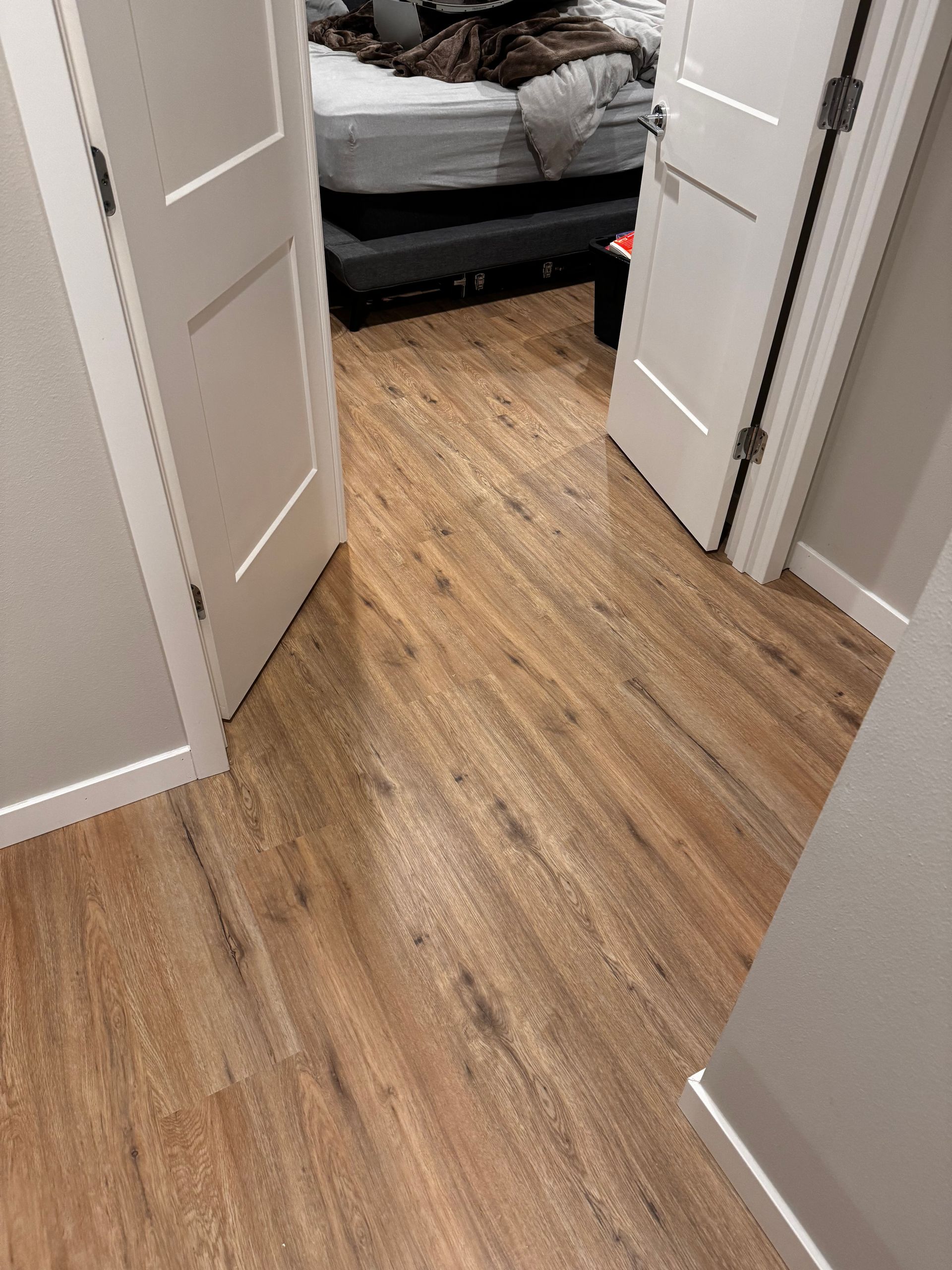 Wood-look flooring in a hallway with open white doors leading to a bed with a rumpled comforter.