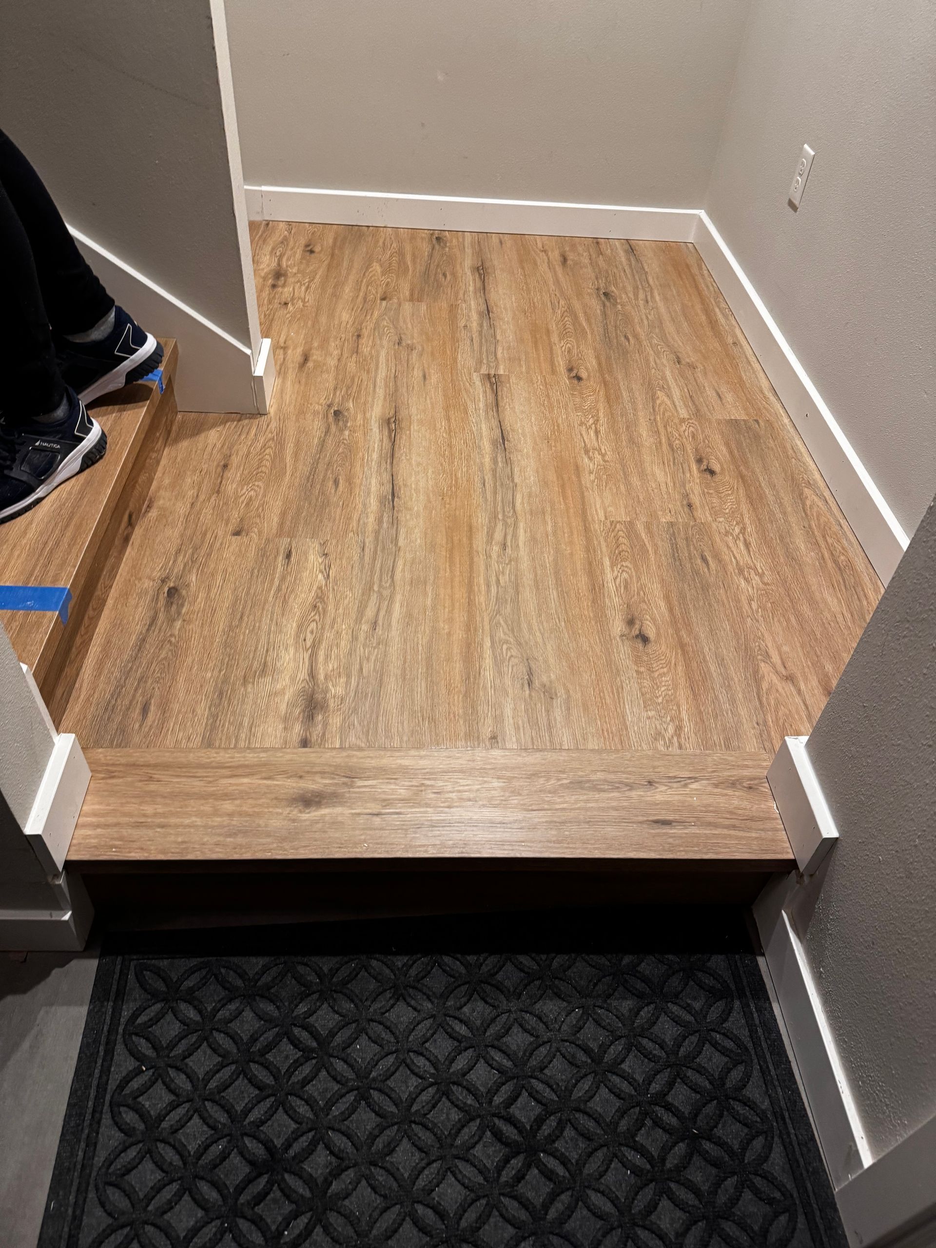 Wooden flooring with white trim in a doorway. A person's legs and shoes are visible on the stairs.