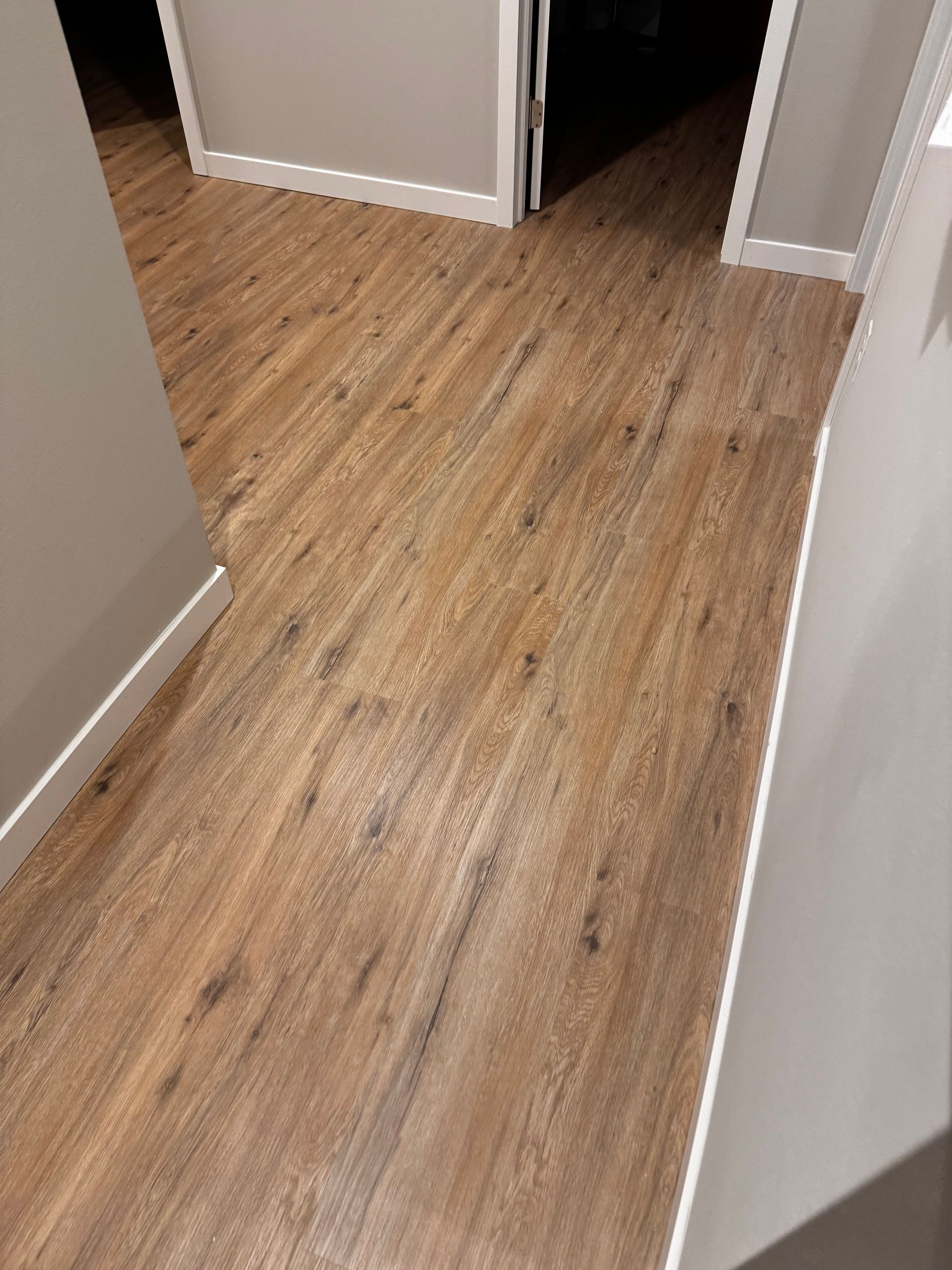 Wooden-look flooring in a hallway with white trim, leading towards an open doorway.