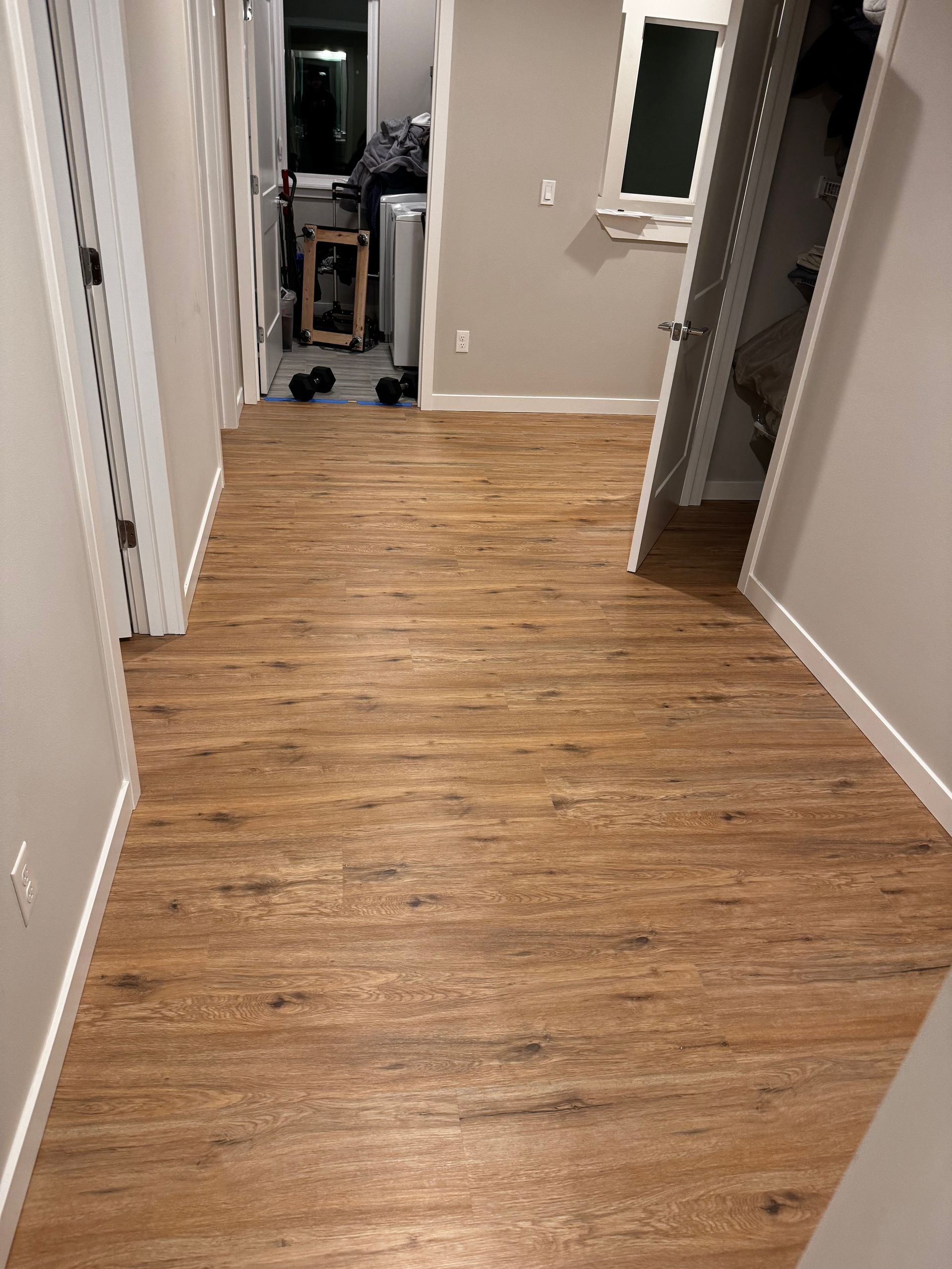 Wood-look flooring in a hallway, leading to a doorway, other rooms visible, white trim, neutral walls.