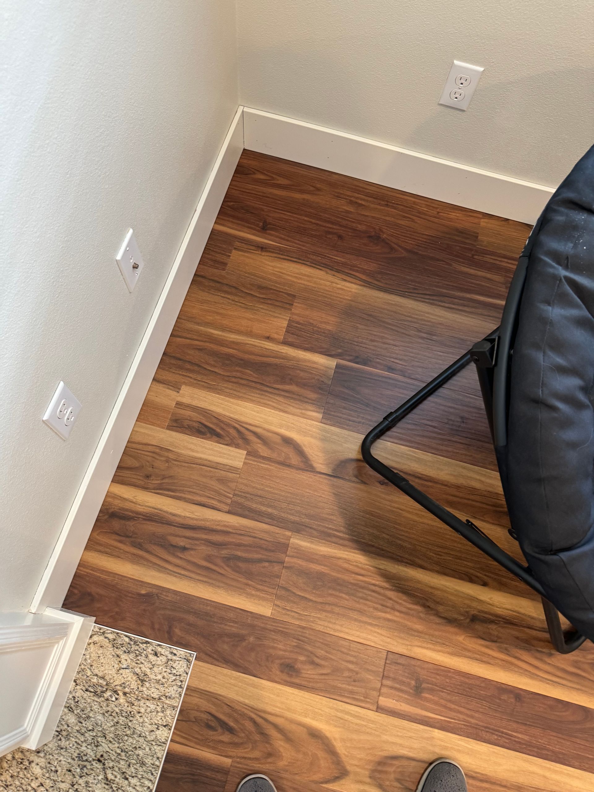 Brown and tan wood floor, white baseboards, and wall with power outlets; a black chair.