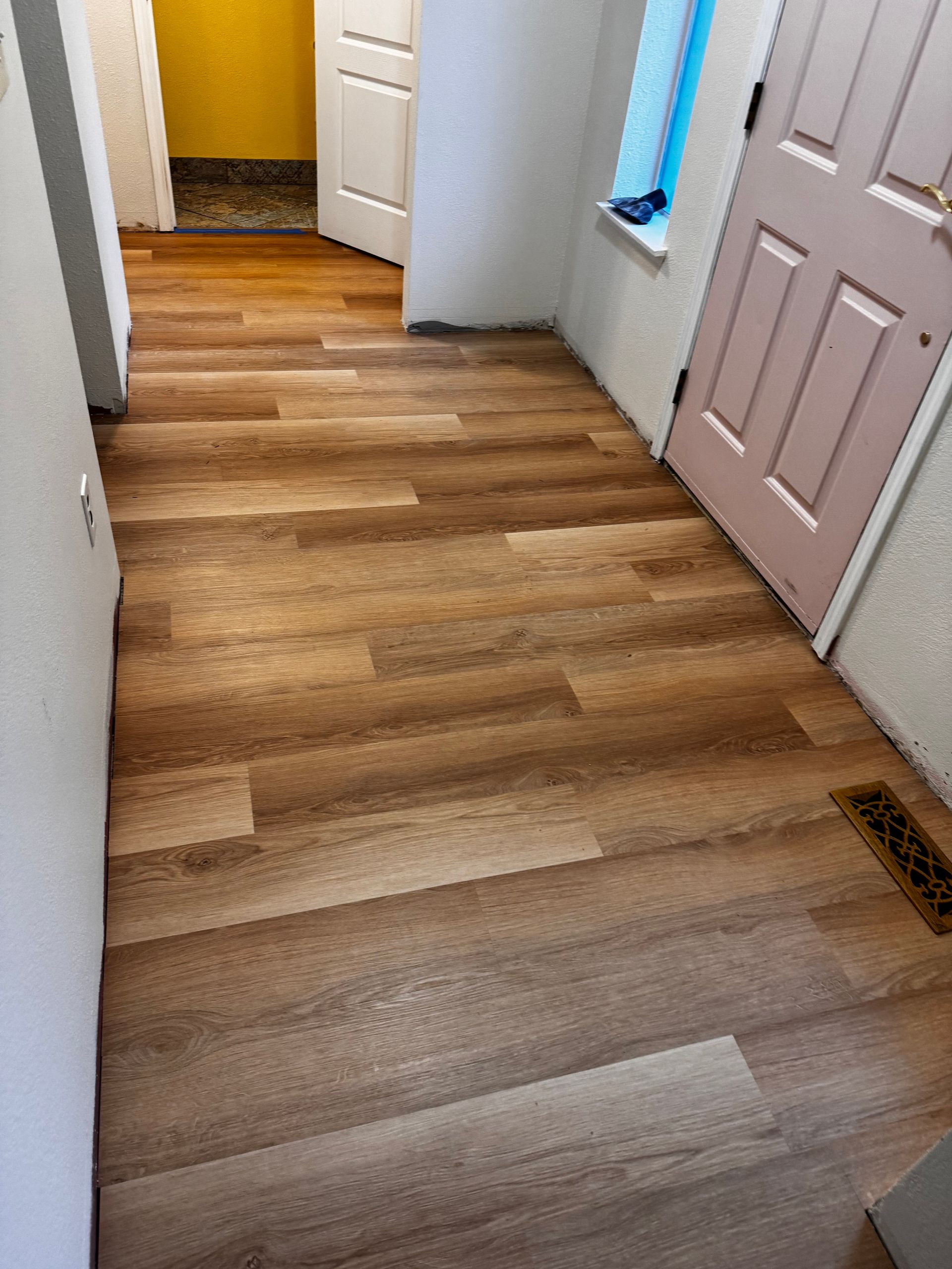 Hallway with wood-look flooring, white walls, and a pink door. Sunlight through a window.