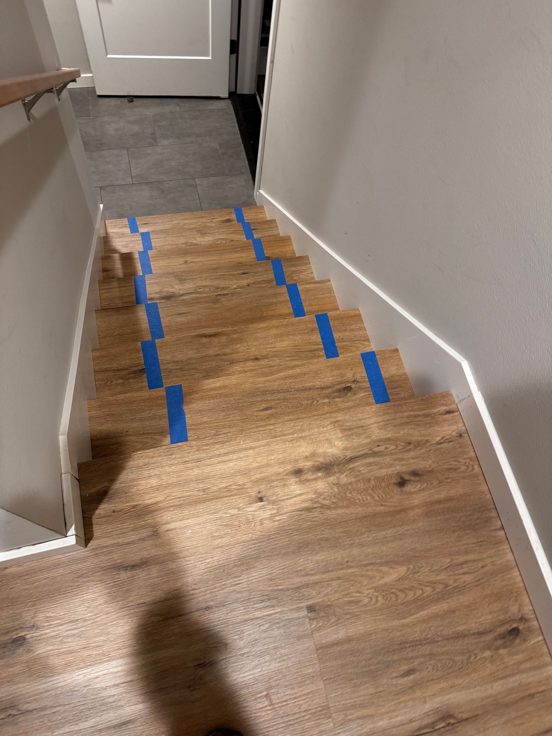 Wooden staircase with blue painter's tape on treads, leading down towards a door and landing.