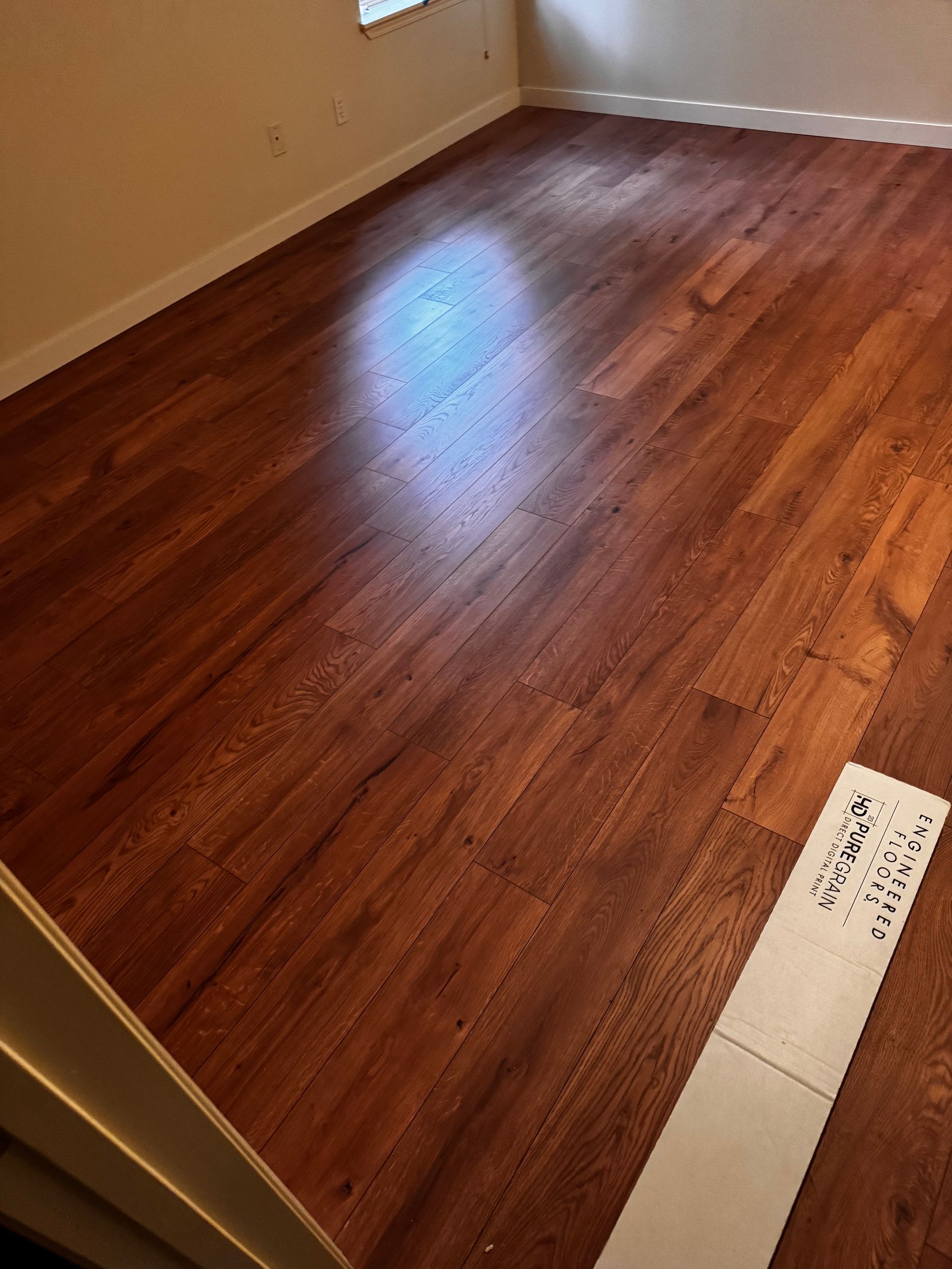 Wooden floor in a room, with sunlight reflecting off its shiny surface. A piece of paper is in the corner.