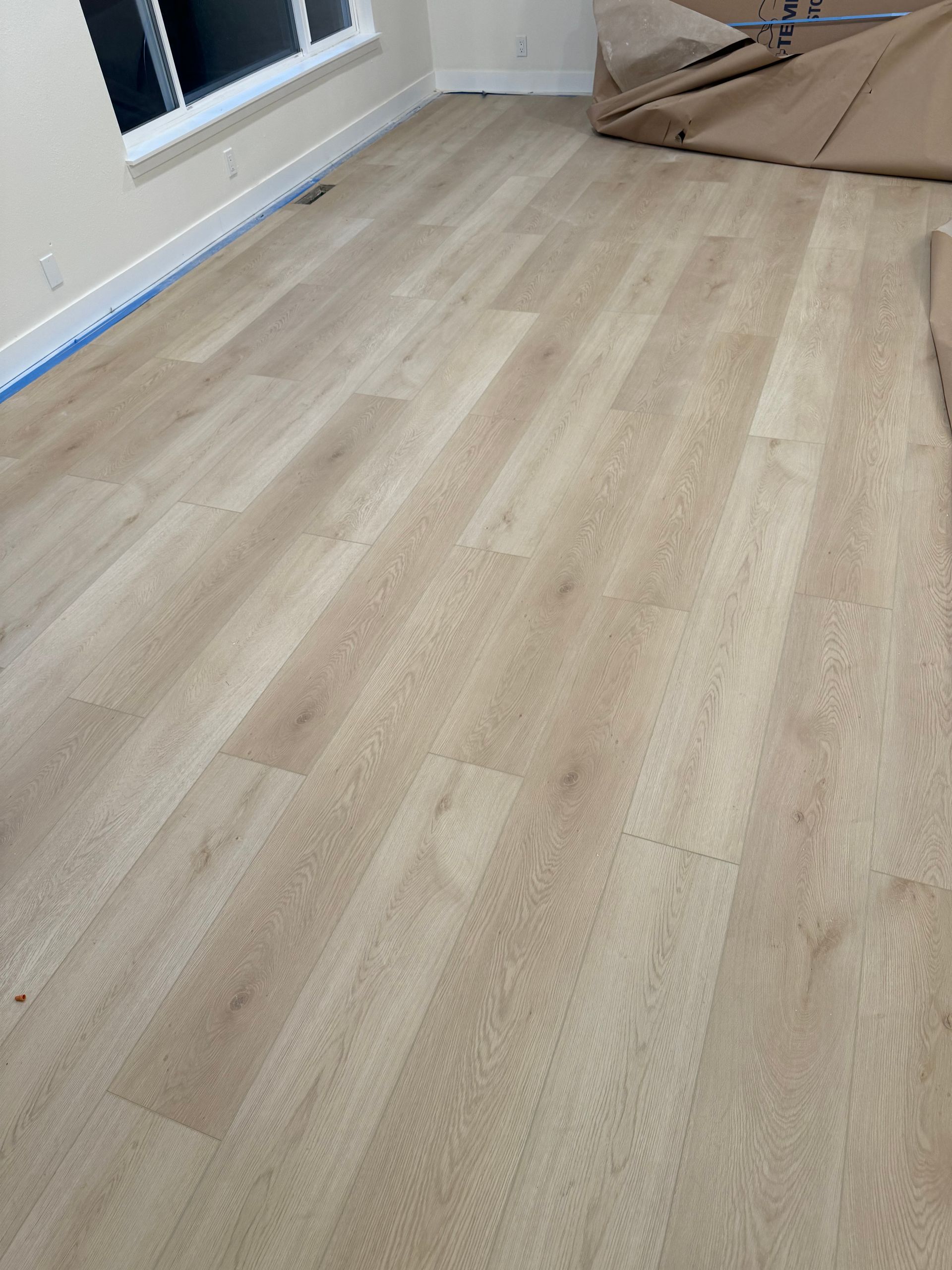 Light-colored wood-look flooring installed in a room. Blue tape lines the baseboards.