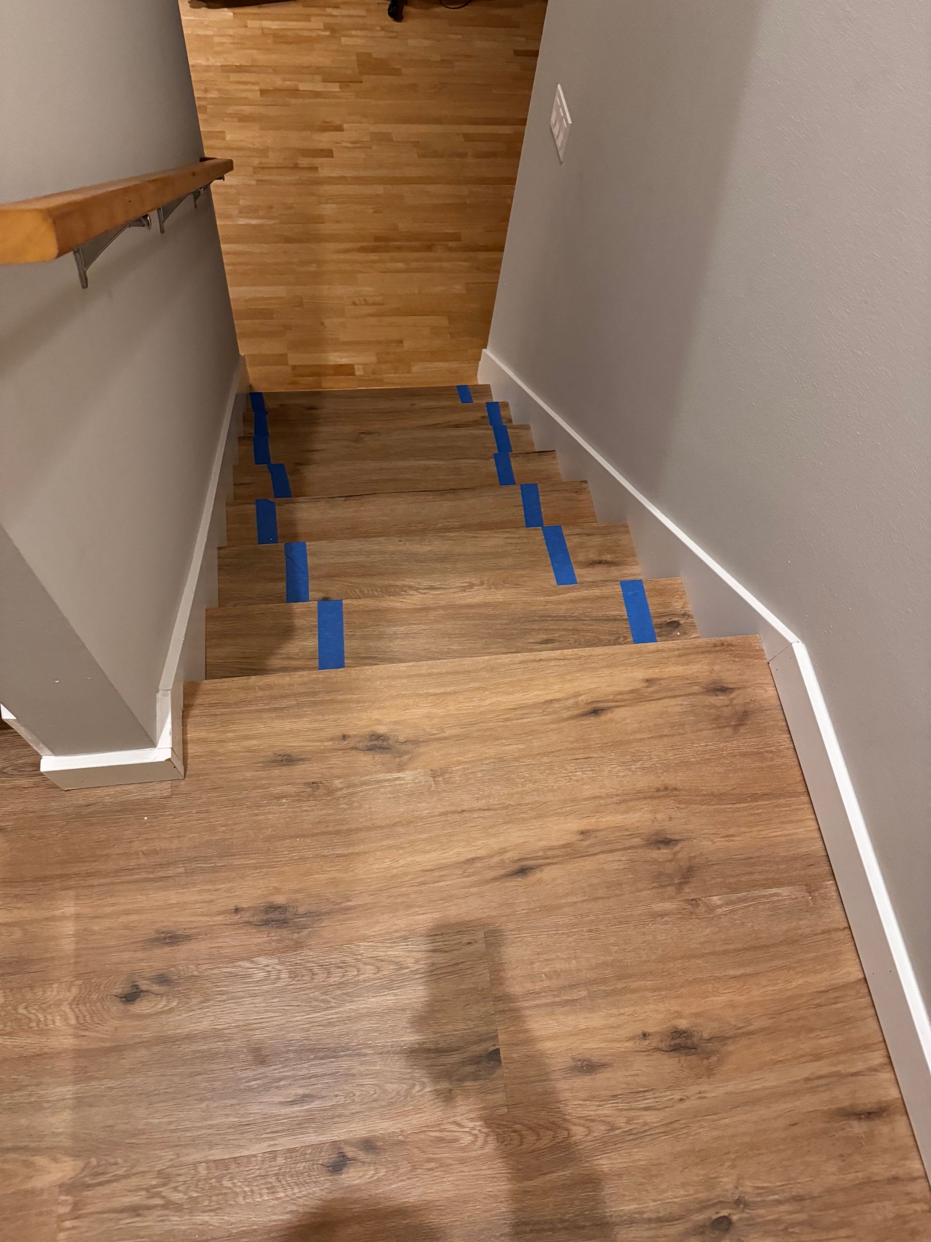 Wooden staircase with blue tape on some steps, between gray walls, and a wooden handrail.