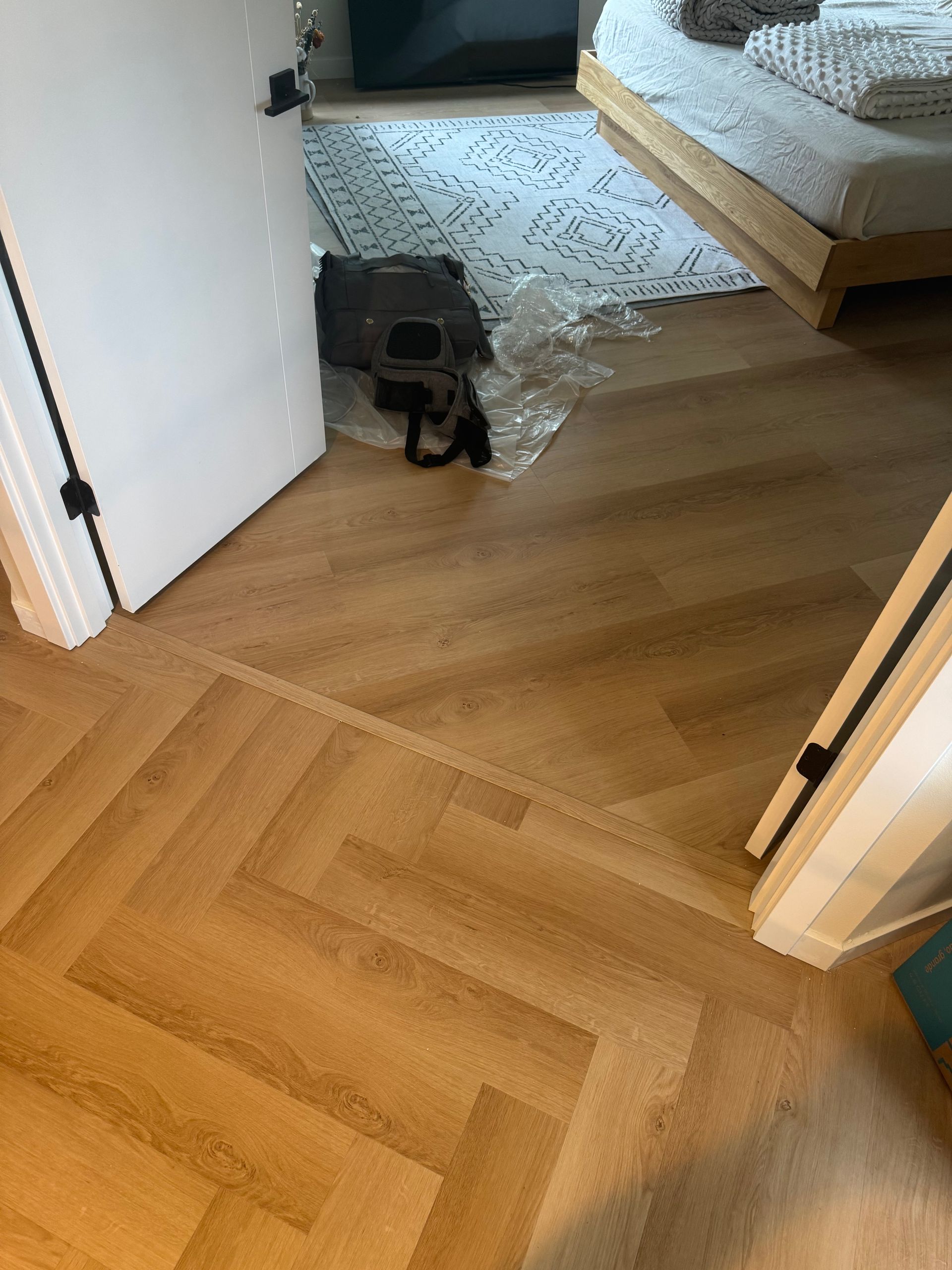 Herringbone patterned wood floor, doorway, and room with bed. A vacuum cleaner and plastic wrapping are in the doorway.