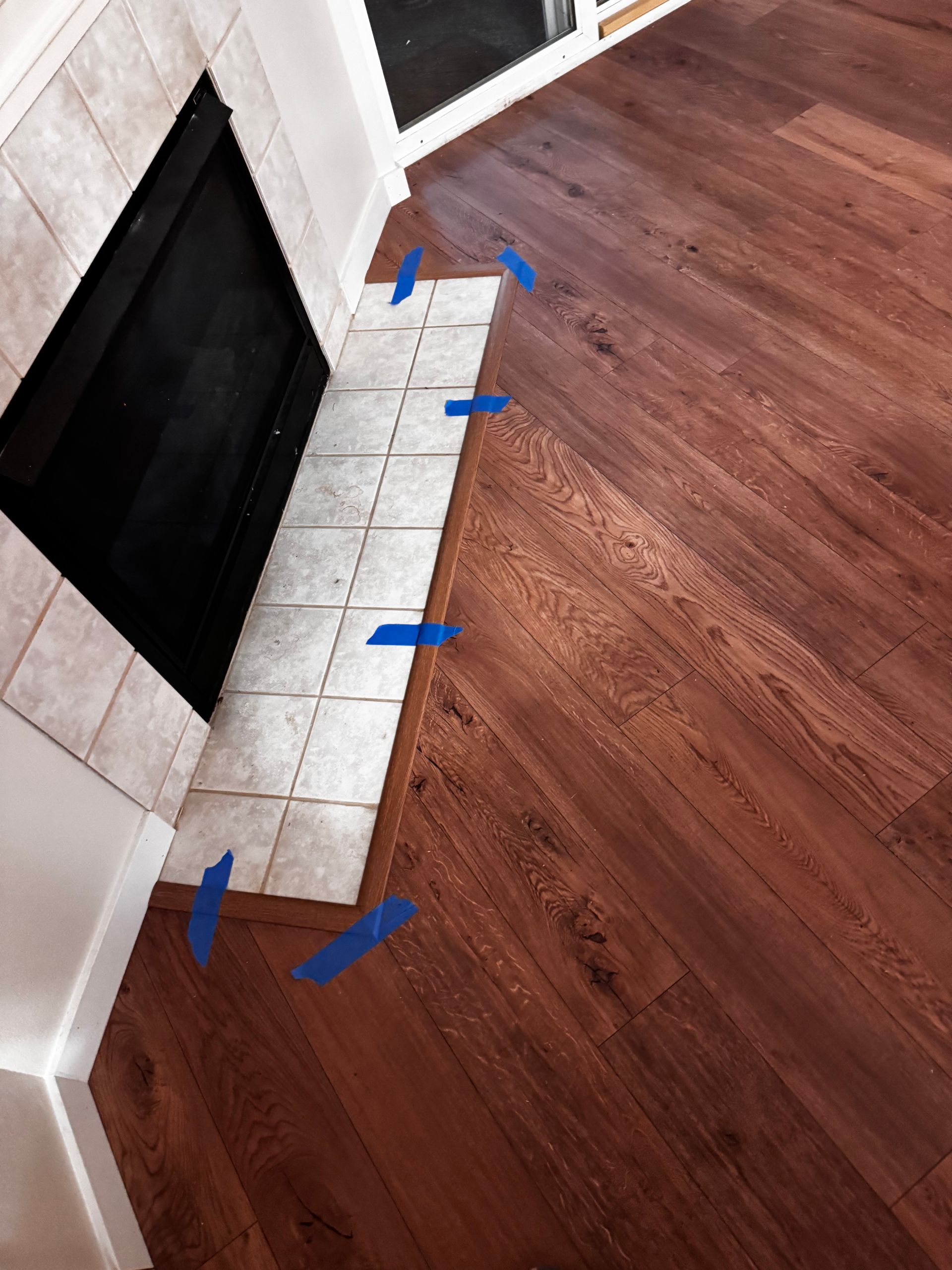 Dark wood-look flooring meets a tiled fireplace hearth; blue tape marks the transition.
