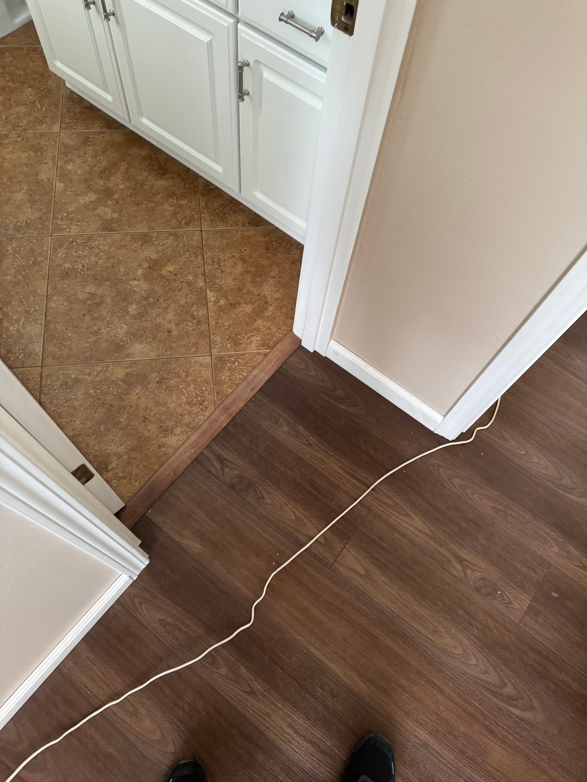 Brown and tan tile floor meets dark wood flooring. White cabinets and a beige wall are present.