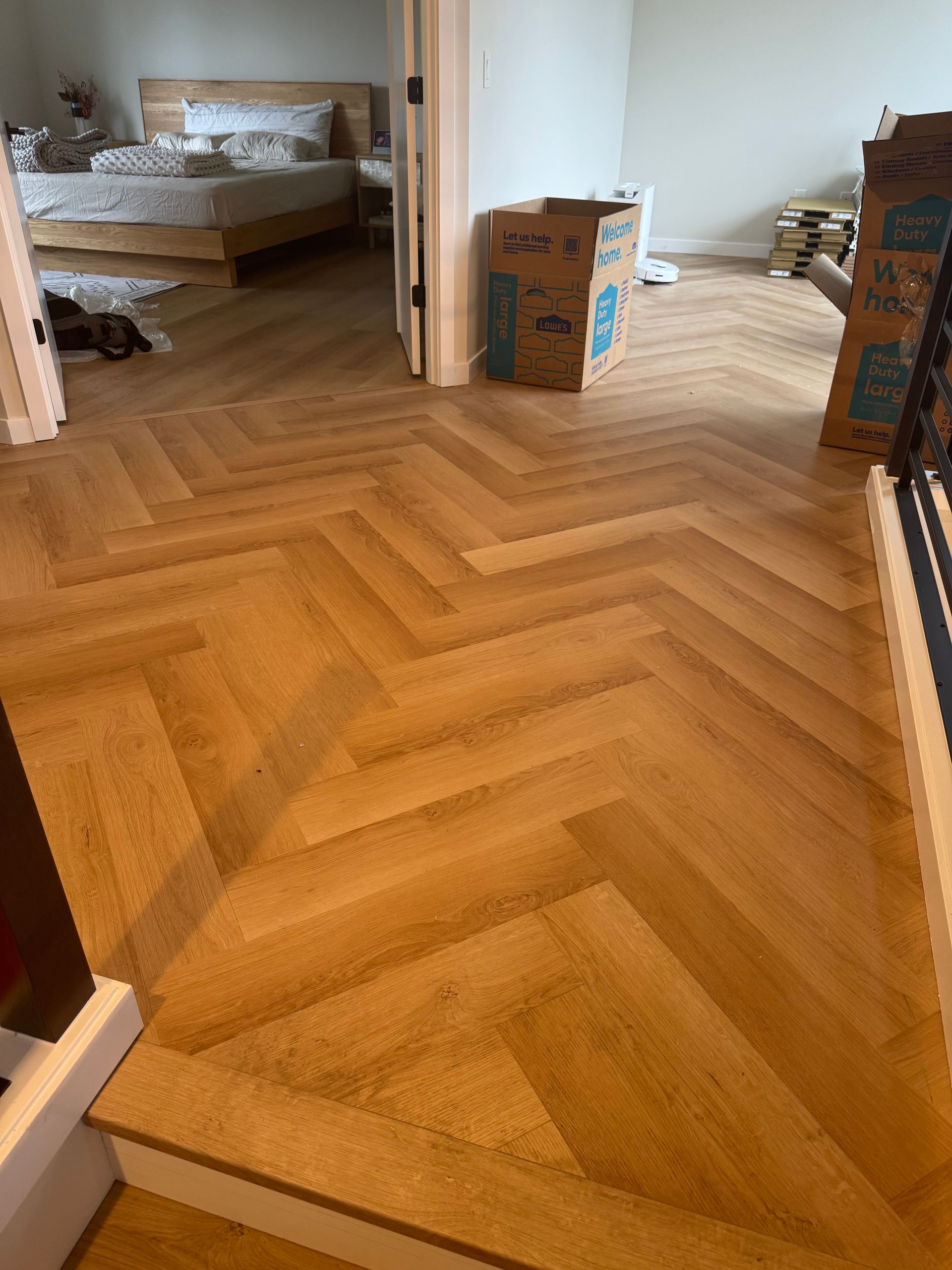 Herringbone wood floor leads to a bedroom with a bed. Boxes are present in the doorway.