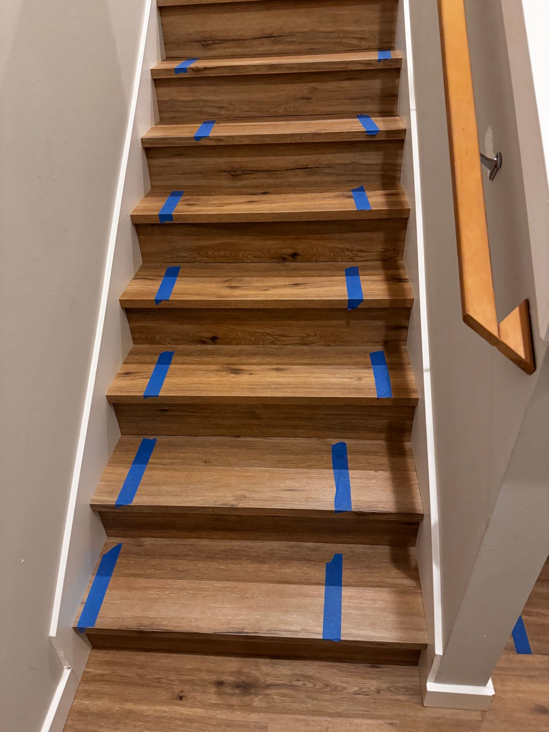 Wooden stairs with blue tape on the treads, beside a white wall and a wooden handrail.
