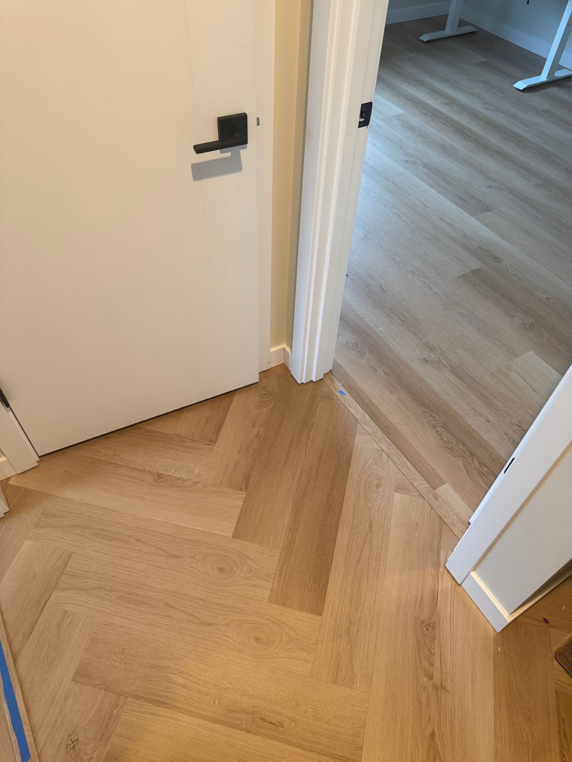 Light-colored herringbone wood floors meet another wood floor in a doorway. White doors and black hardware.