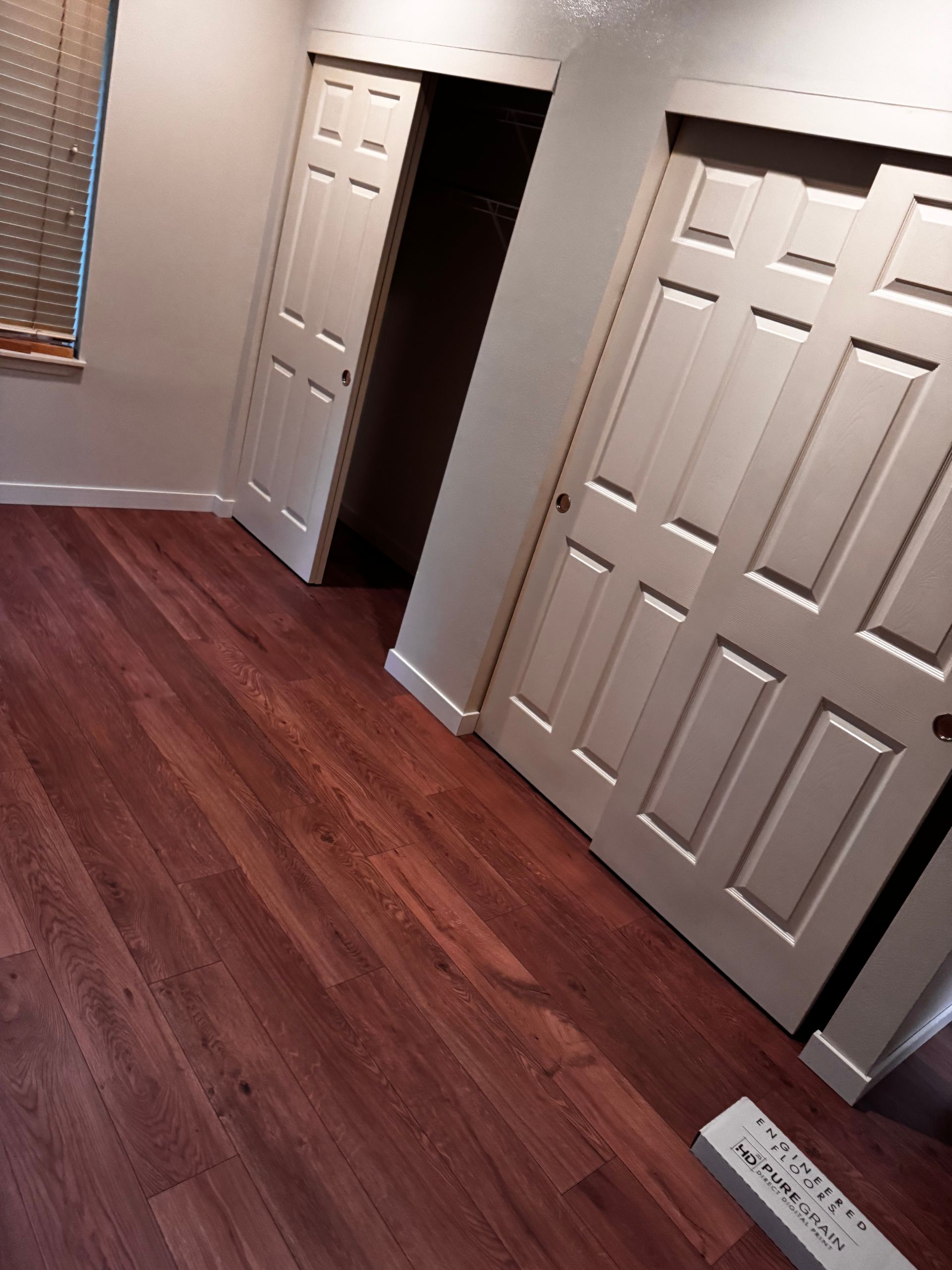 Room with hardwood floors, two white closet doors, and a window with blinds.