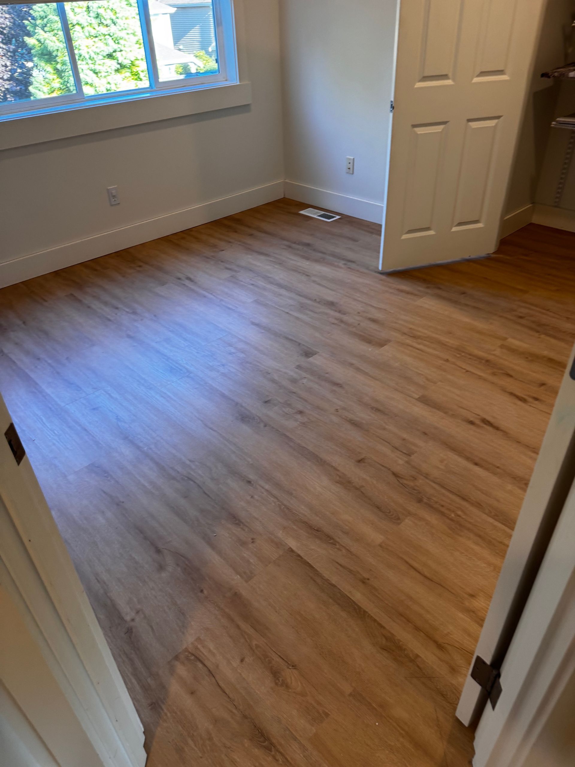 Empty room with wood-look flooring, white trim and door. A window is in the background.