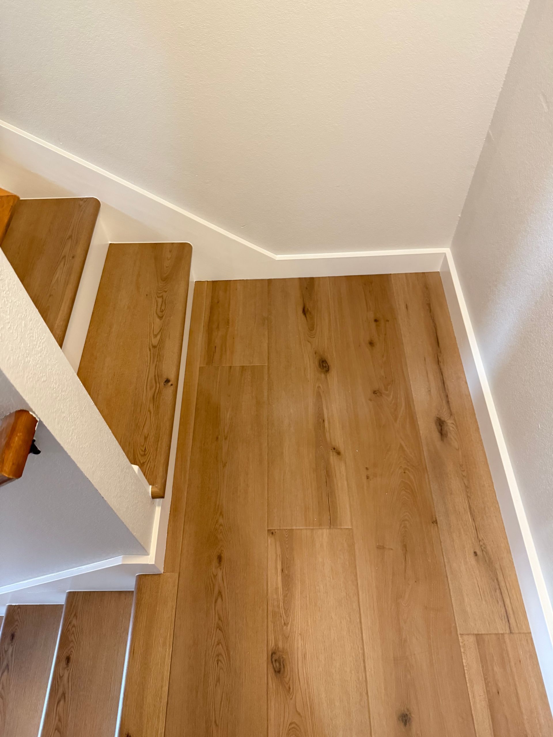 Wooden flooring meets staircase, white trim on wall and stairs.