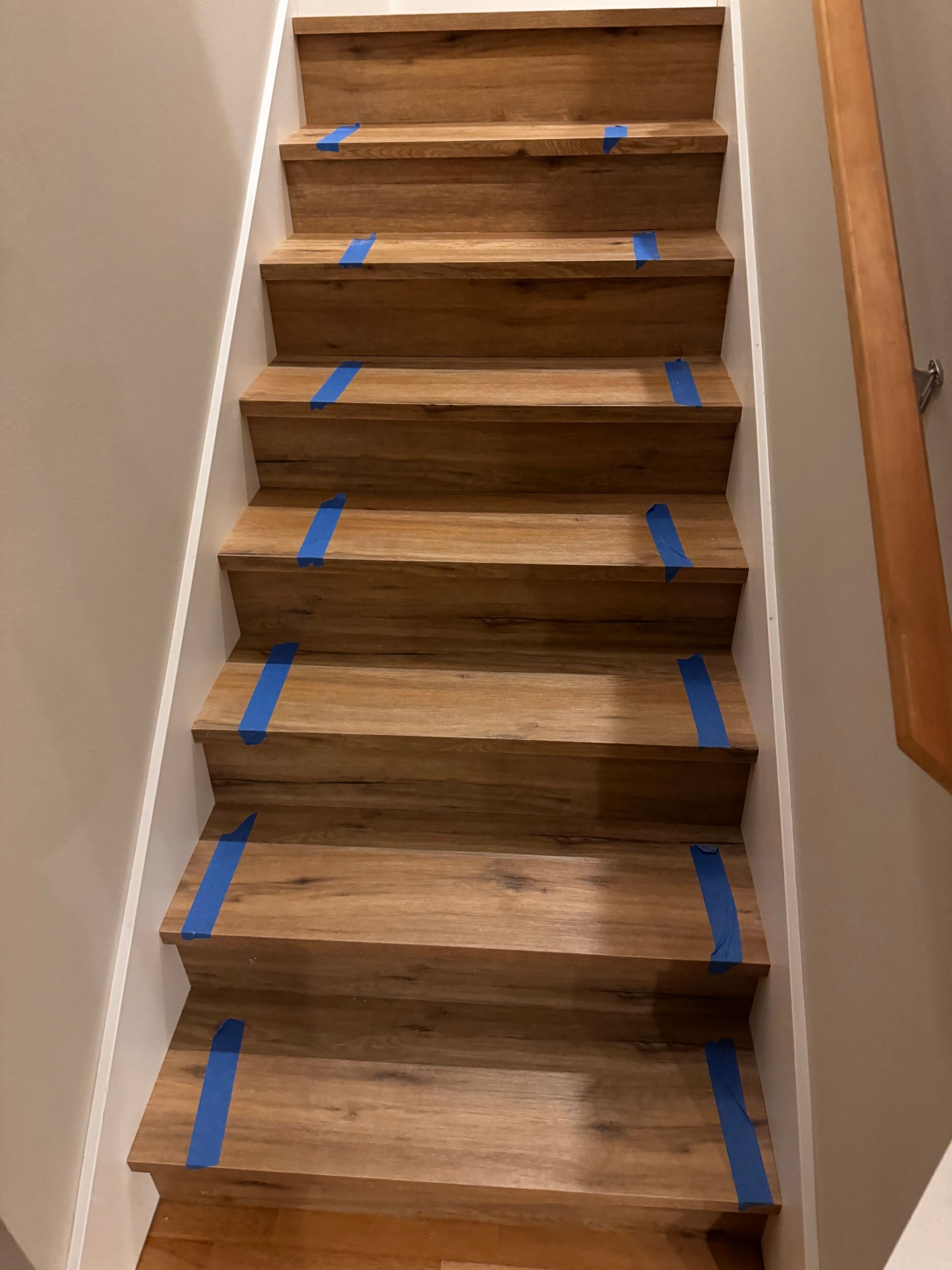 Wooden staircase with blue tape on treads. Beige walls and a wooden handrail are visible.