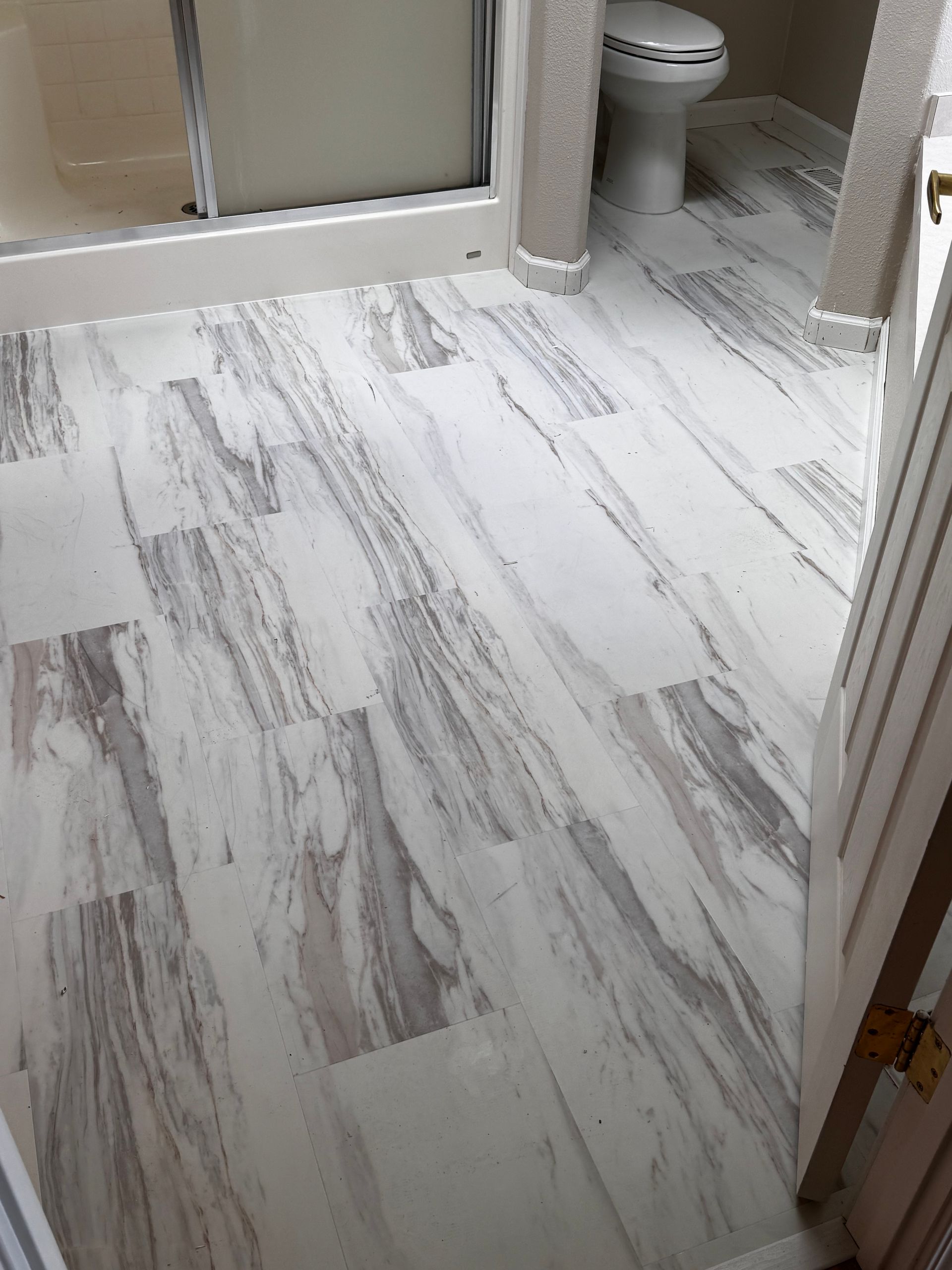 Bathroom with marble-patterned flooring, toilet, shower, and partially visible door. Gray and white tones.