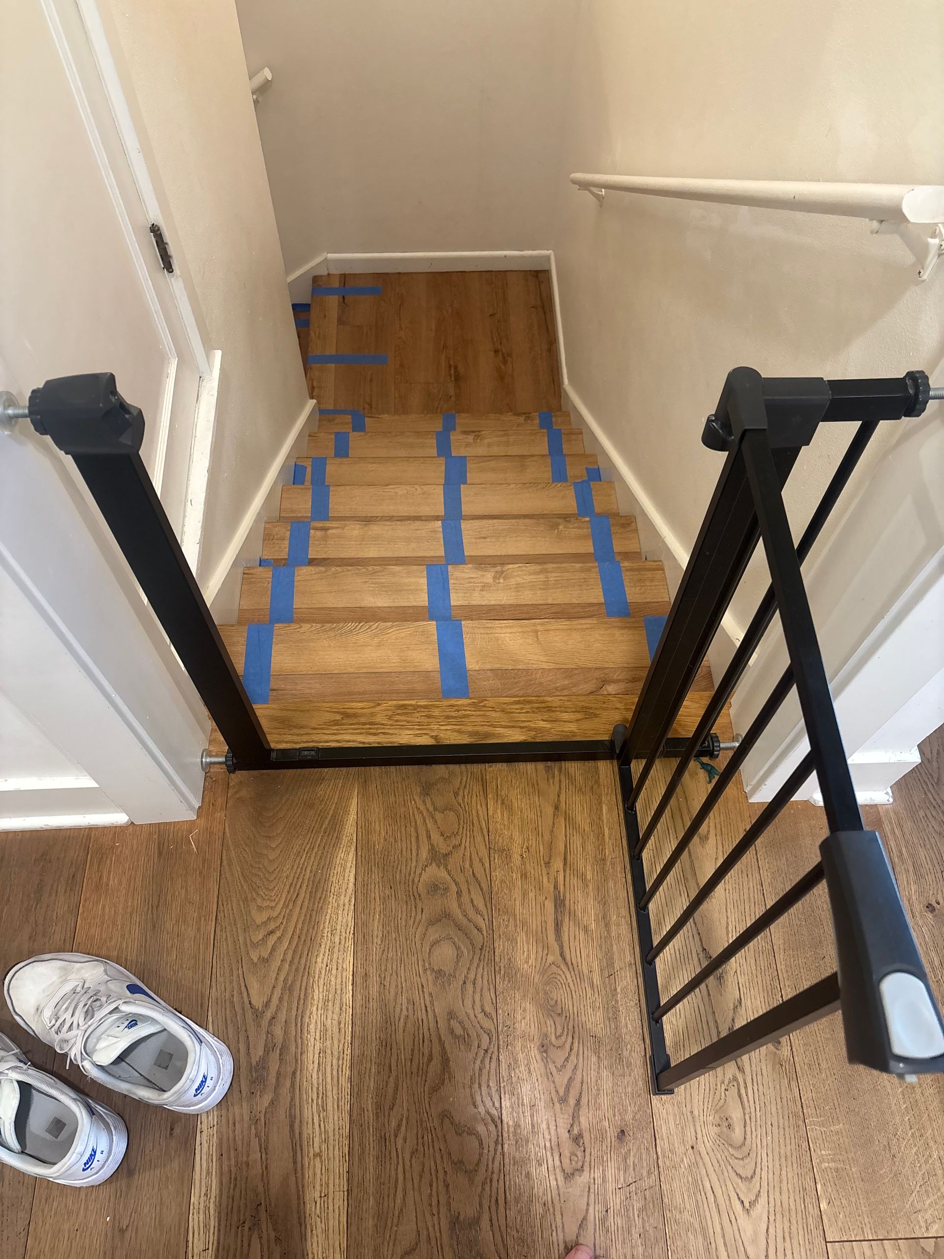 View down a wooden staircase with blue painter's tape on steps and a black baby gate installed.