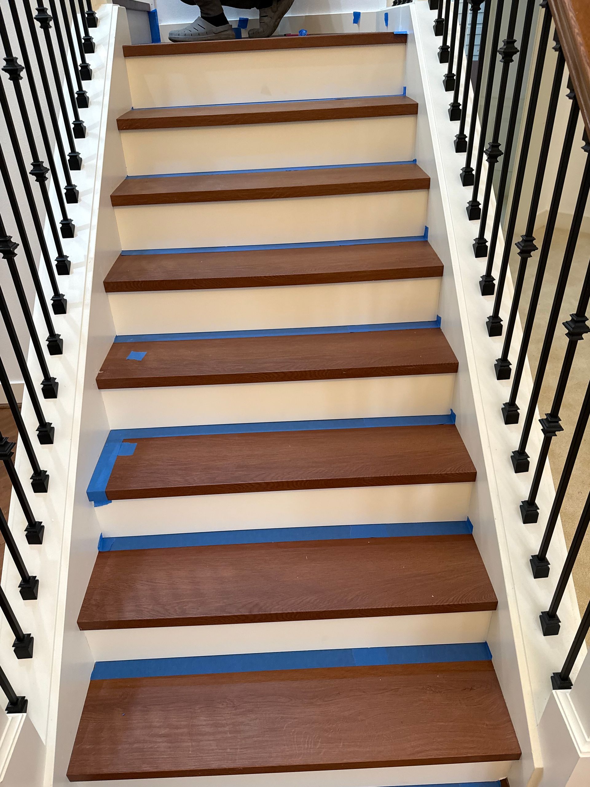 Staircase with brown wooden steps, white risers, and black iron railings. Blue tape is applied.