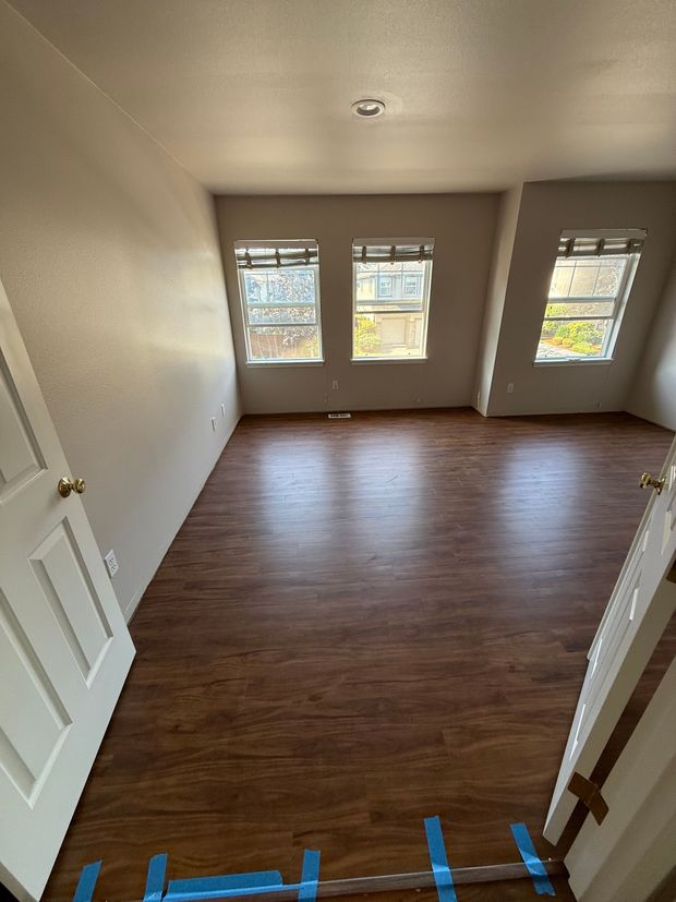 Empty room with wood-look flooring, three windows, and a white door. The walls are light beige.