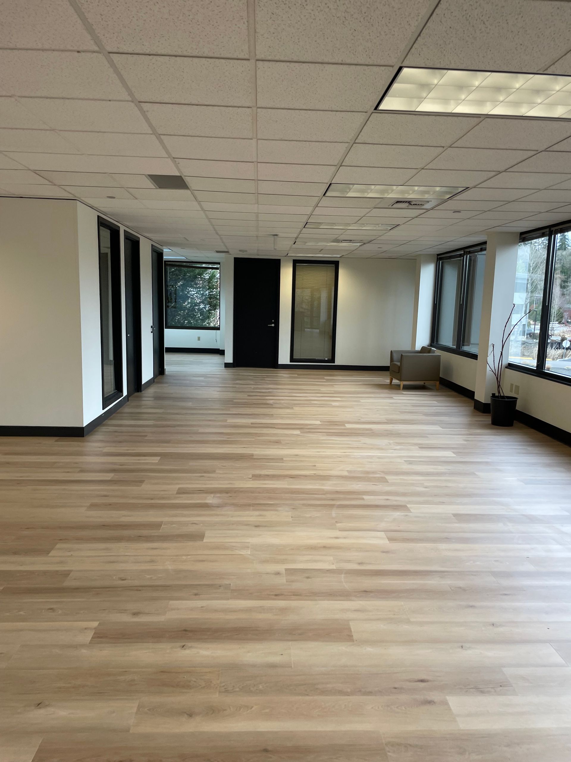 Empty office space with light wood-look flooring, white walls, and large windows.
