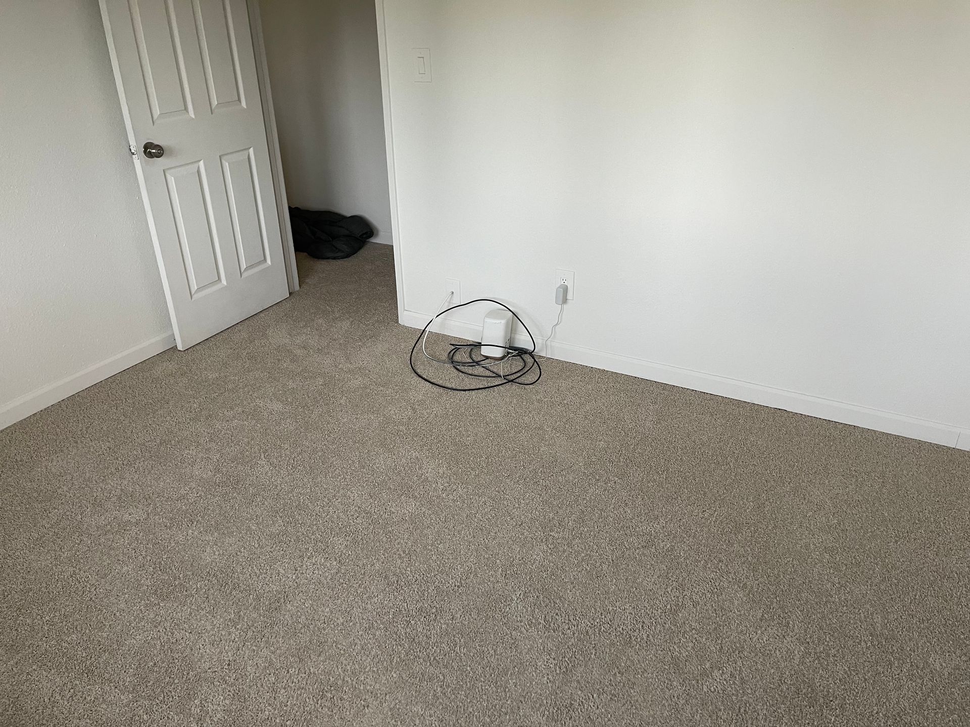 Empty room with beige carpet, white walls, and a closed door. Cables on the floor.