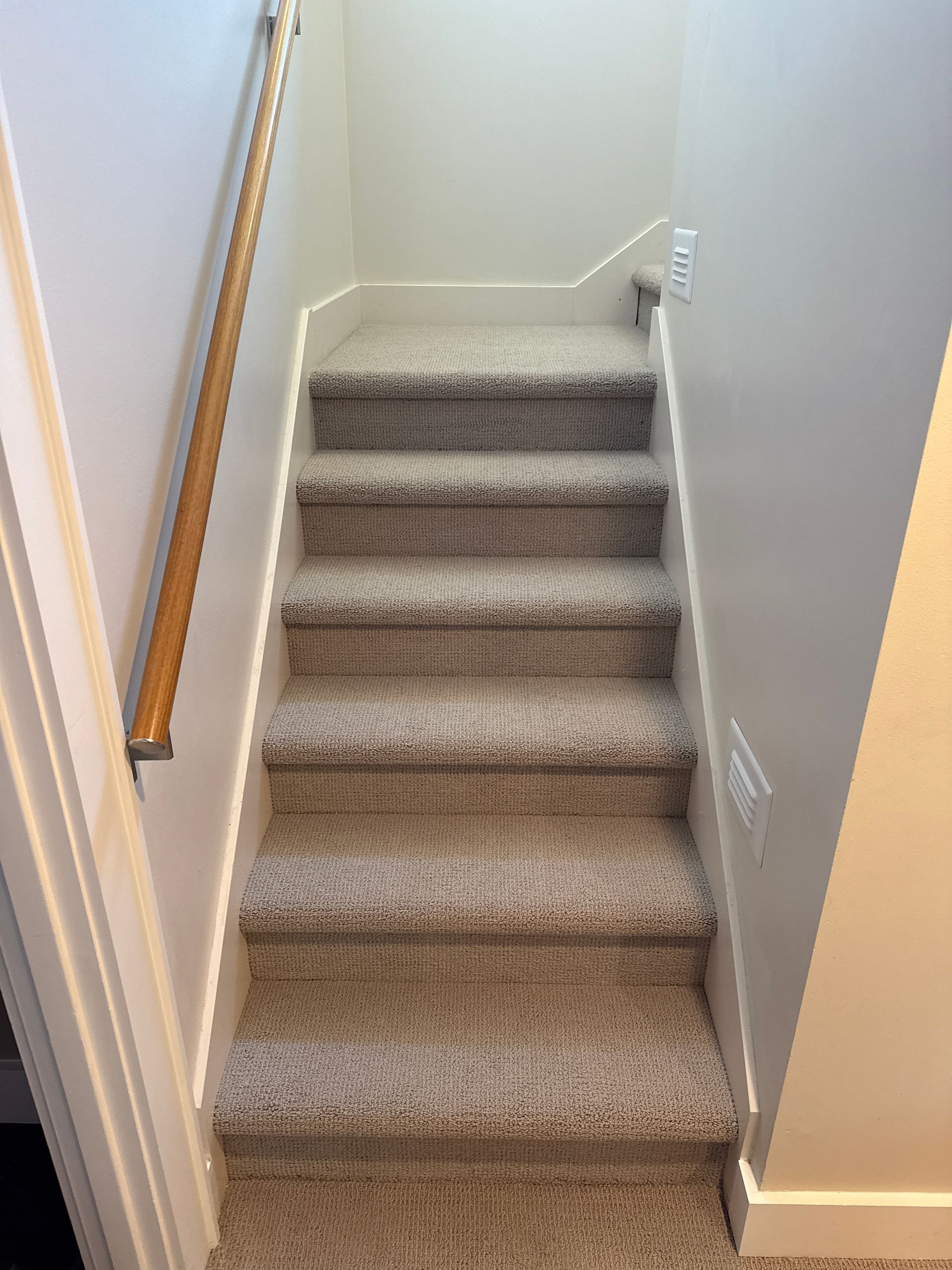Carpeted staircase with a wooden handrail, leading upwards, set between two light walls.