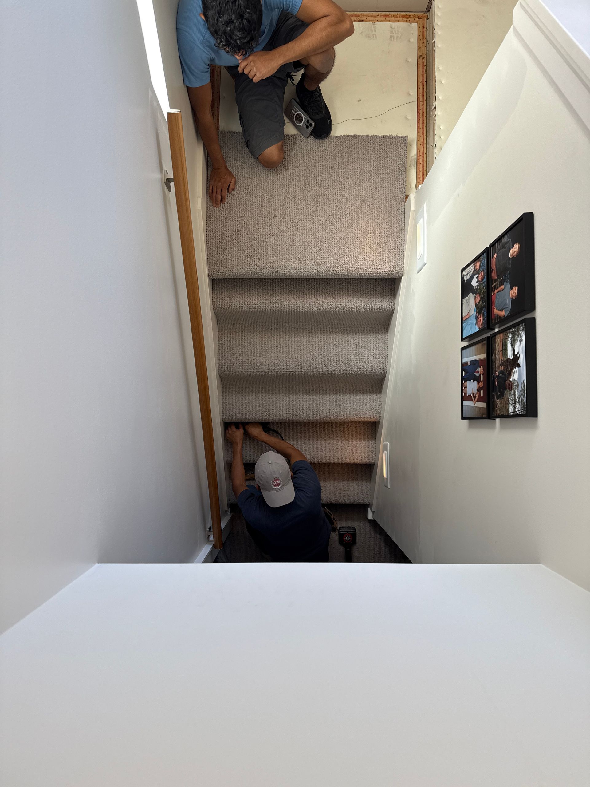 Two people installing carpet on a staircase. One works below, the other above. White walls surround them.