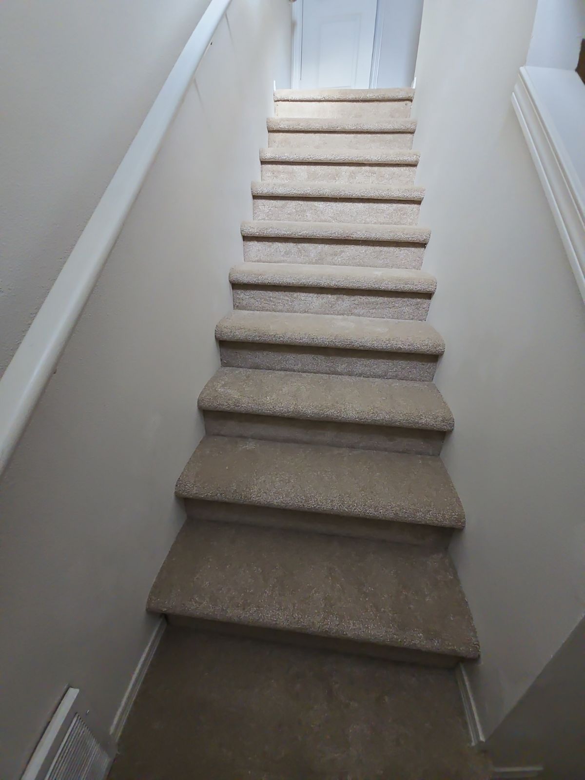 Staircase with carpeted steps descends between beige walls. White handrail on the left.