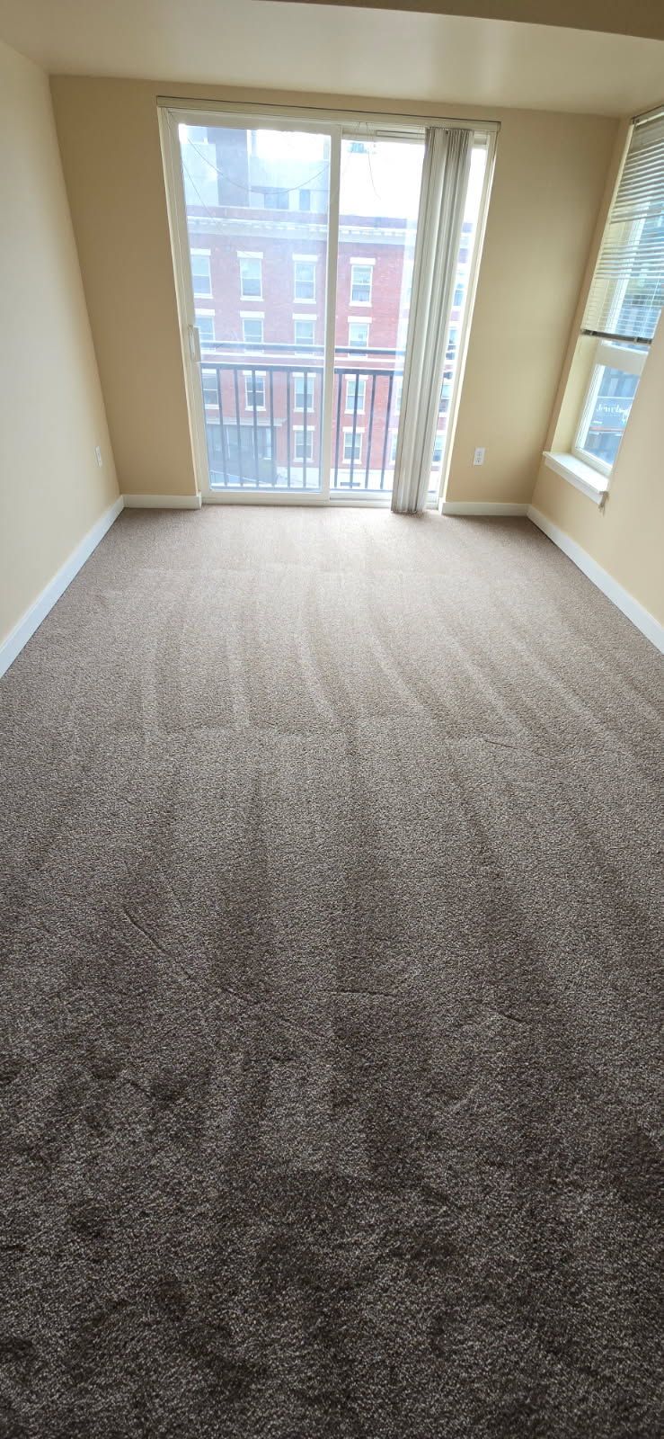 An empty room with brown carpet, sliding glass door leading to a balcony, and a window.