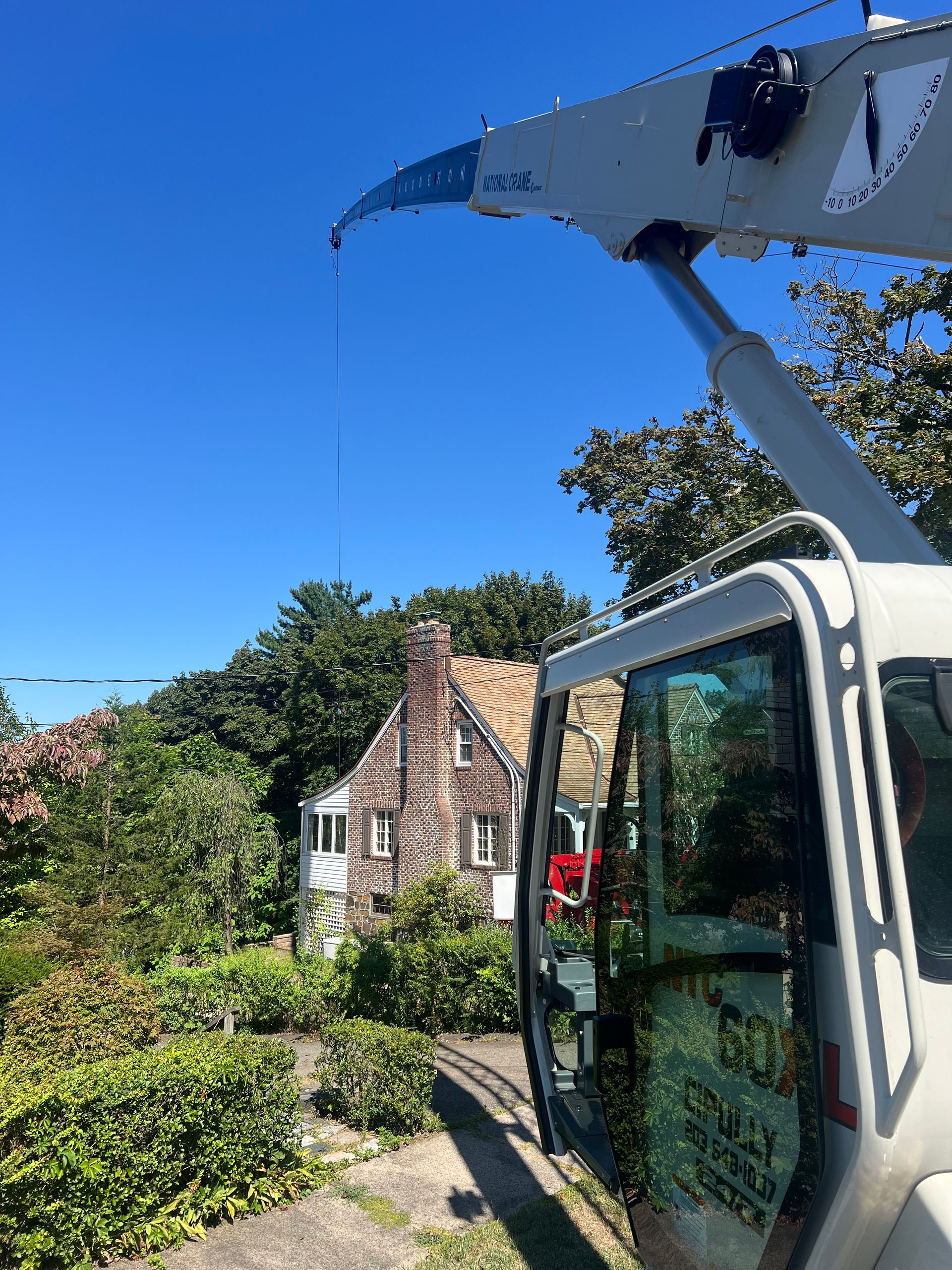 Crane boom and cab foreground; house behind trees, clear blue sky.
