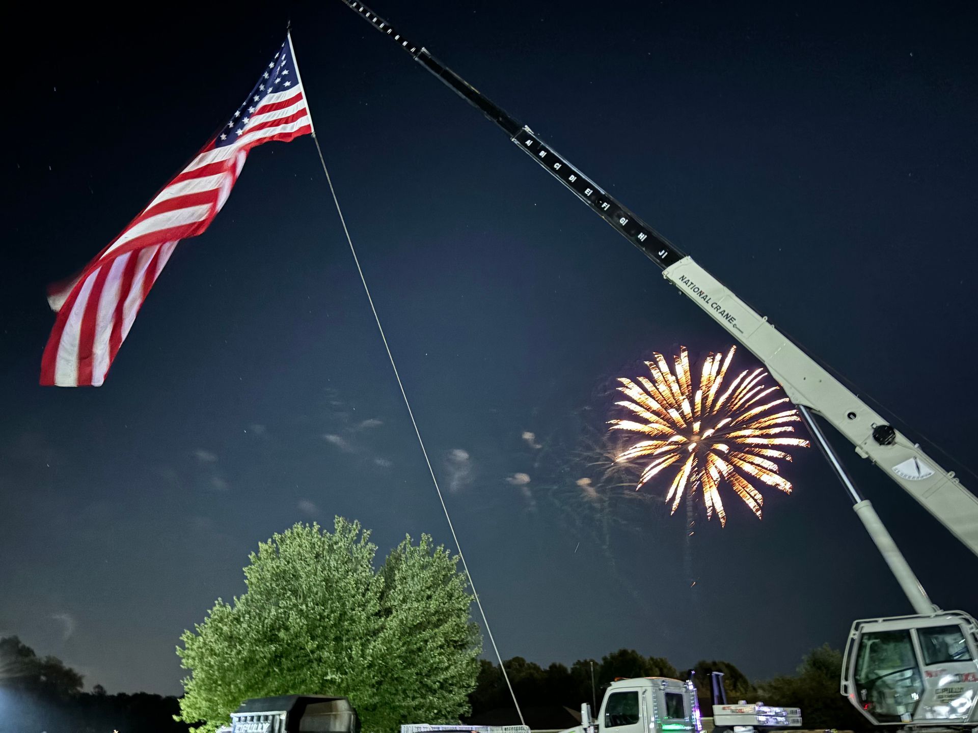 A crane is lifting an american flag in front of a fireworks display.