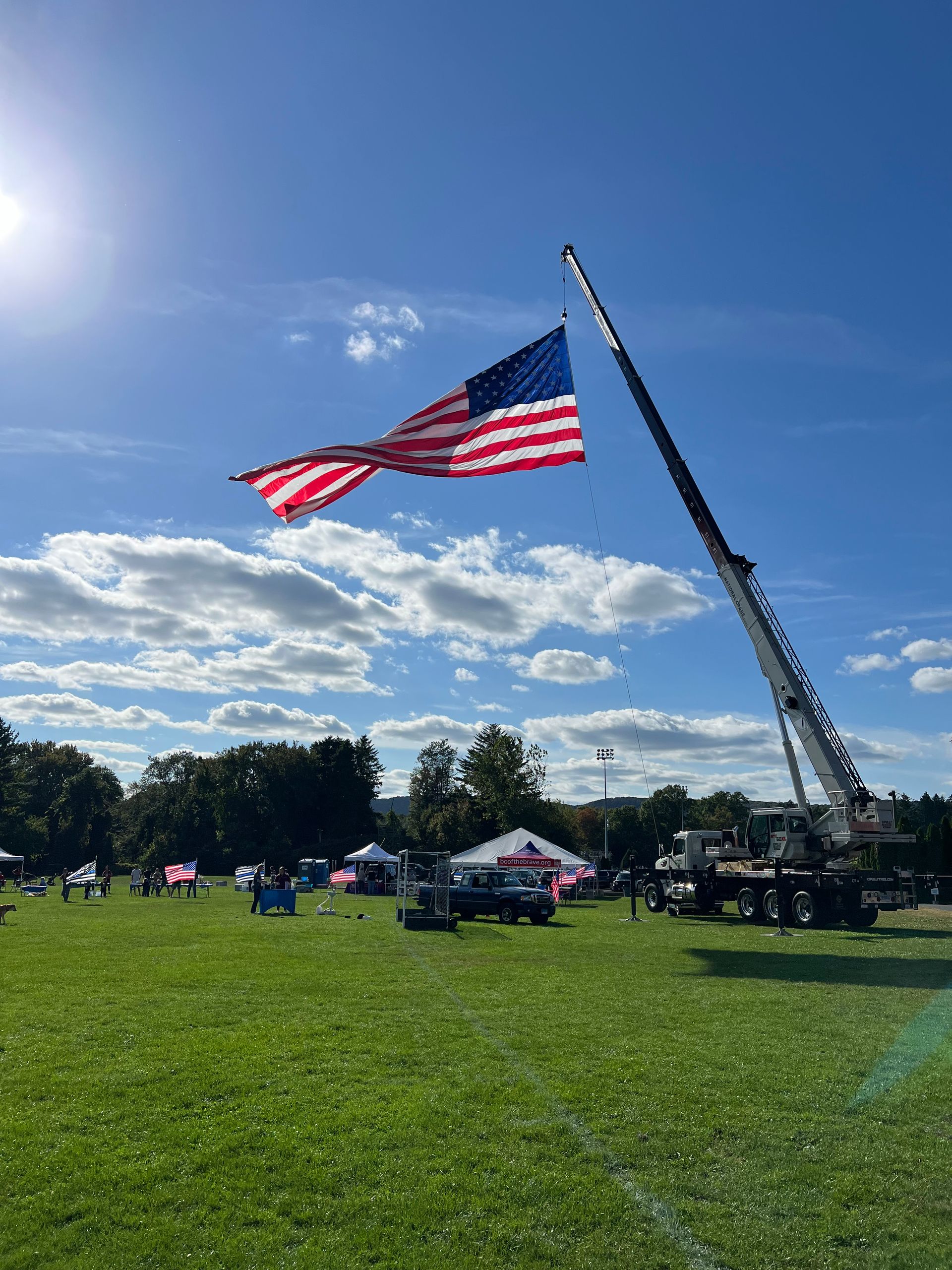 A large american flag is being lifted by a crane in a grassy field.