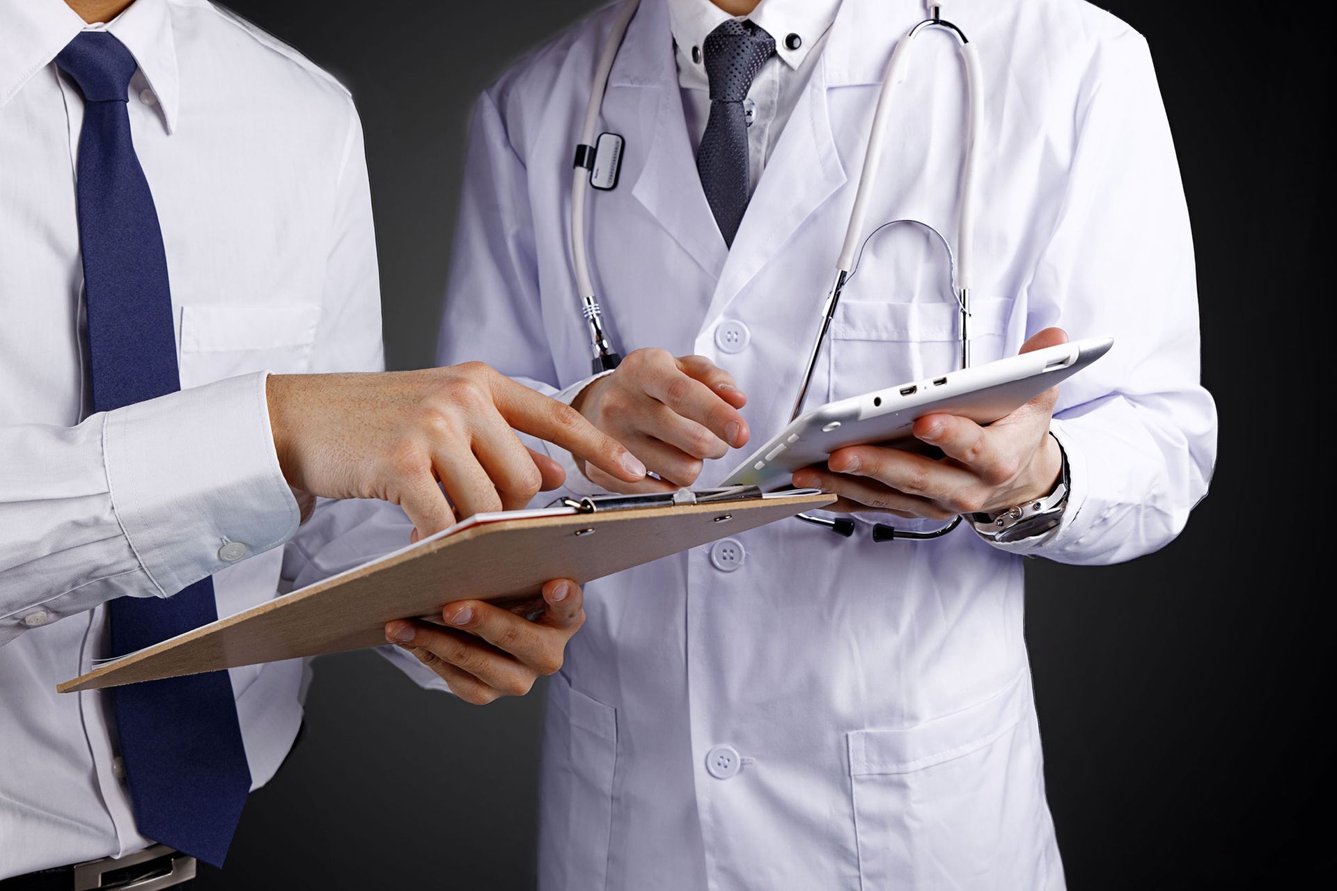 Two people in white coats, one with a clipboard, the other with a tablet, discussing data.