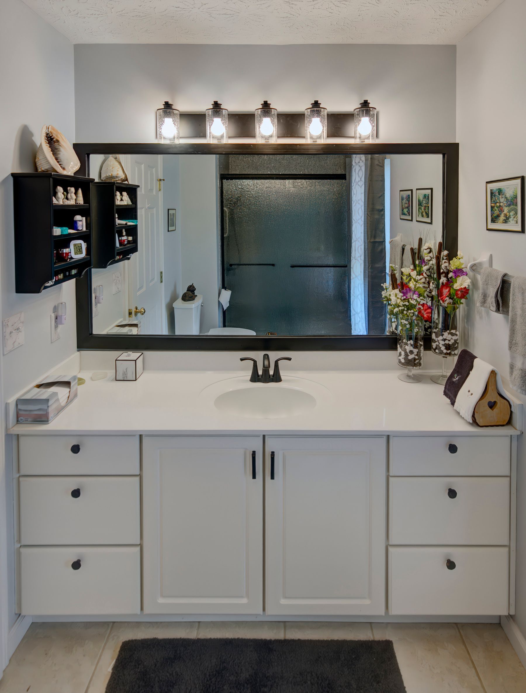 Bathroom with white vanity, large mirror, and black accents. Gray walls, dark gray rug, and a small black storage cabinet.