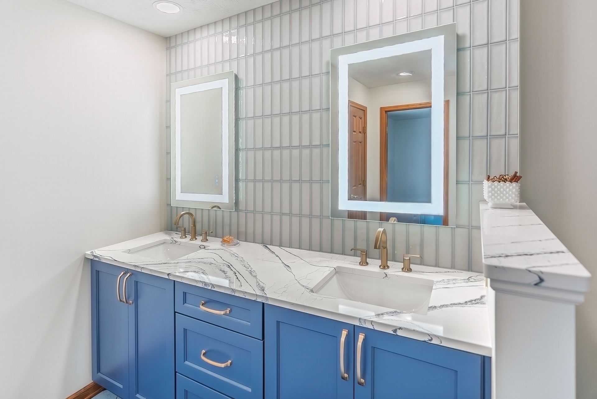 Blue bathroom vanity with marble countertop and two sinks, mirrors with lights, and gray tiled wall.