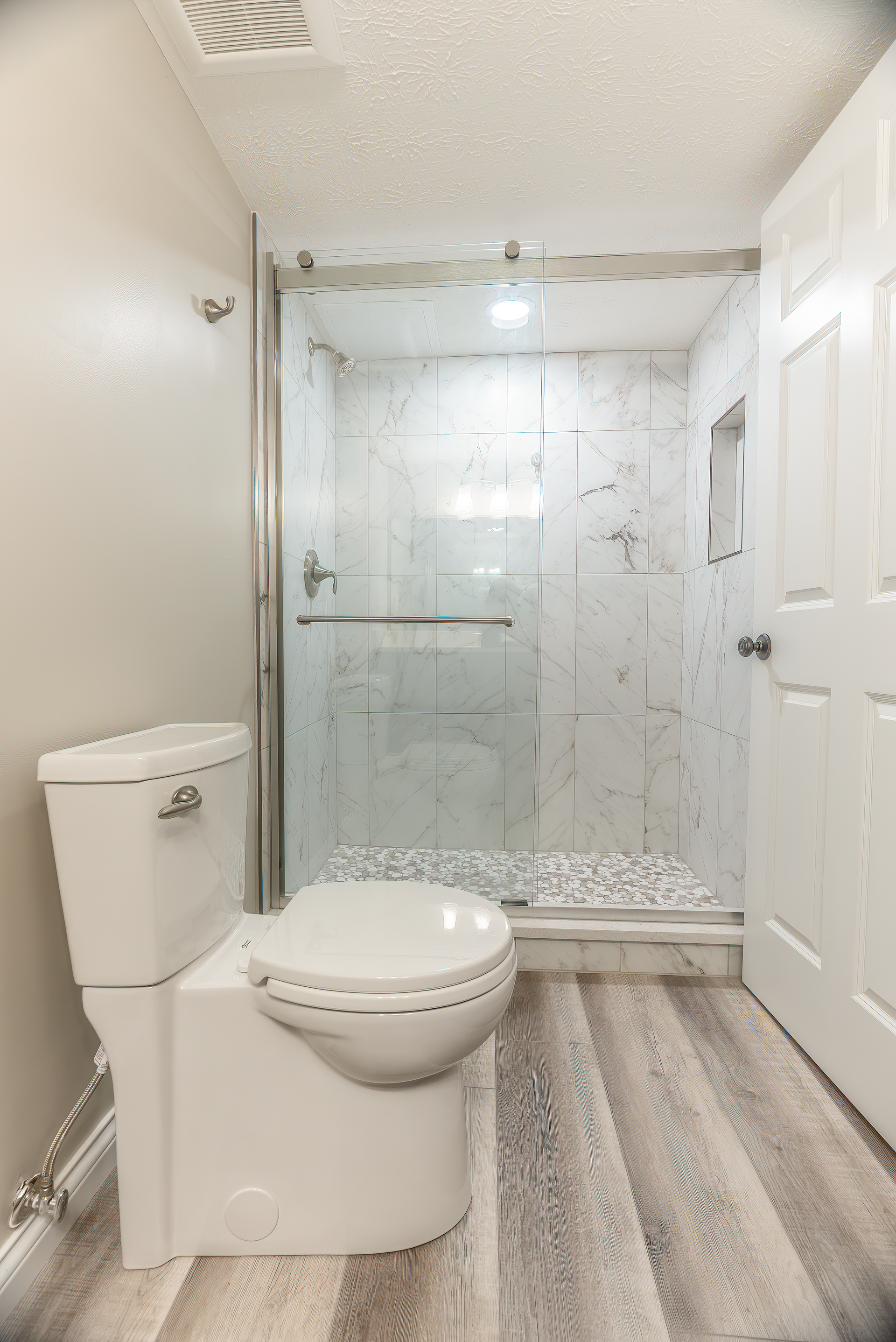 Modern bathroom with white toilet, marble-tiled shower, glass door, and gray wood-look flooring.