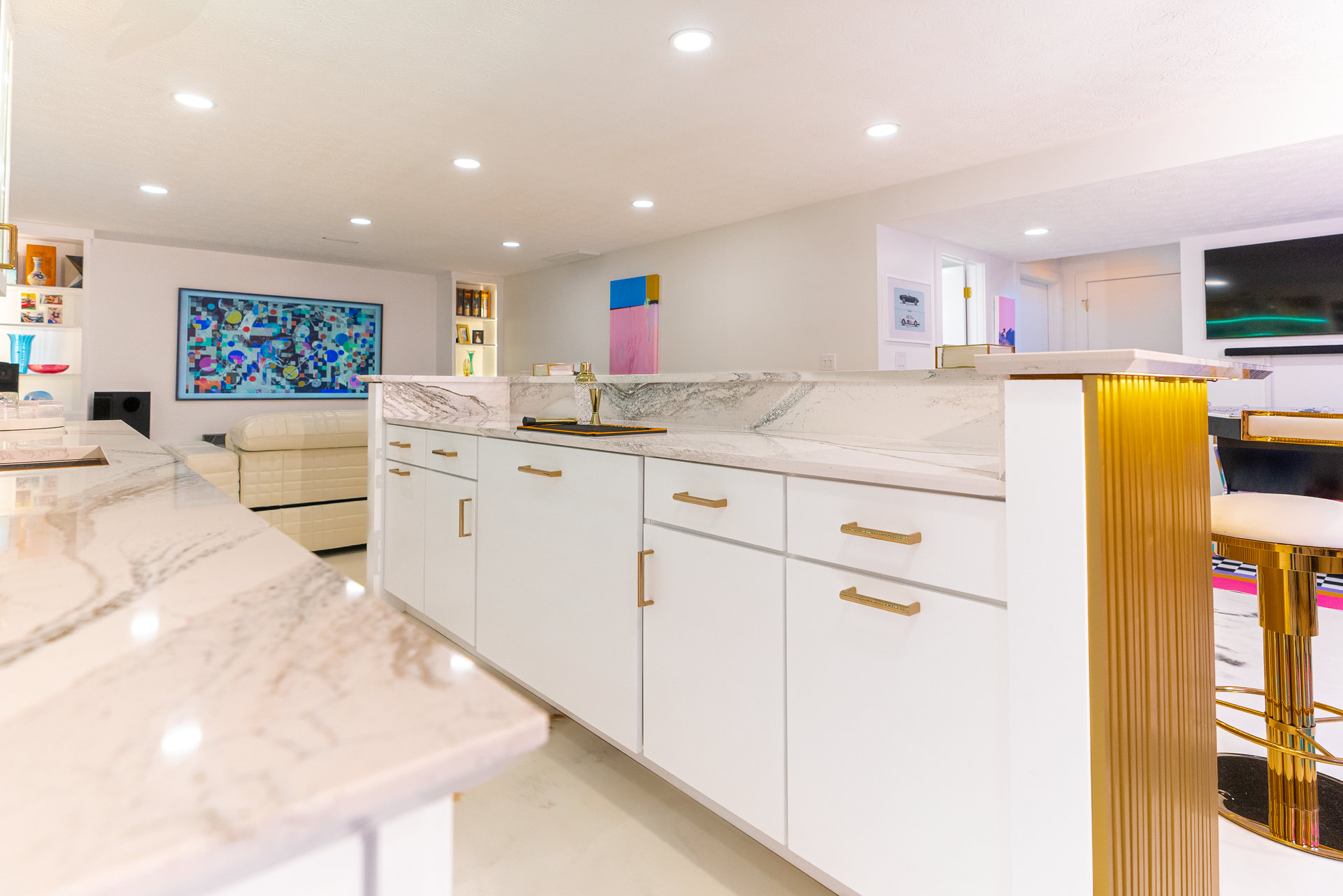 A bright modern kitchen with white cabinetry, gold accents, marble countertops, and a colorful abstract painting.