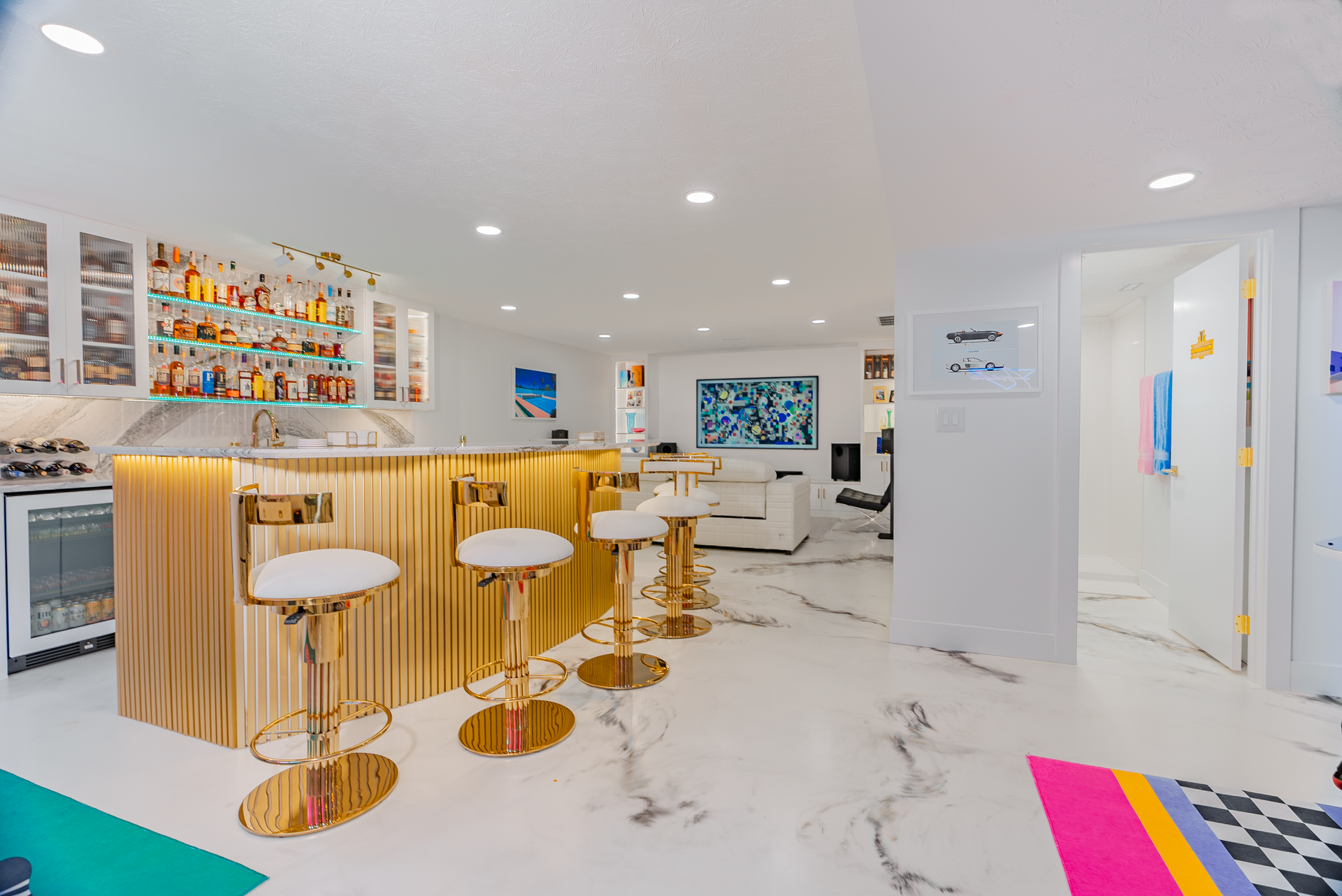 Gold bar with white stools in a bright room with art, a colorful rug, and a small refrigerator.