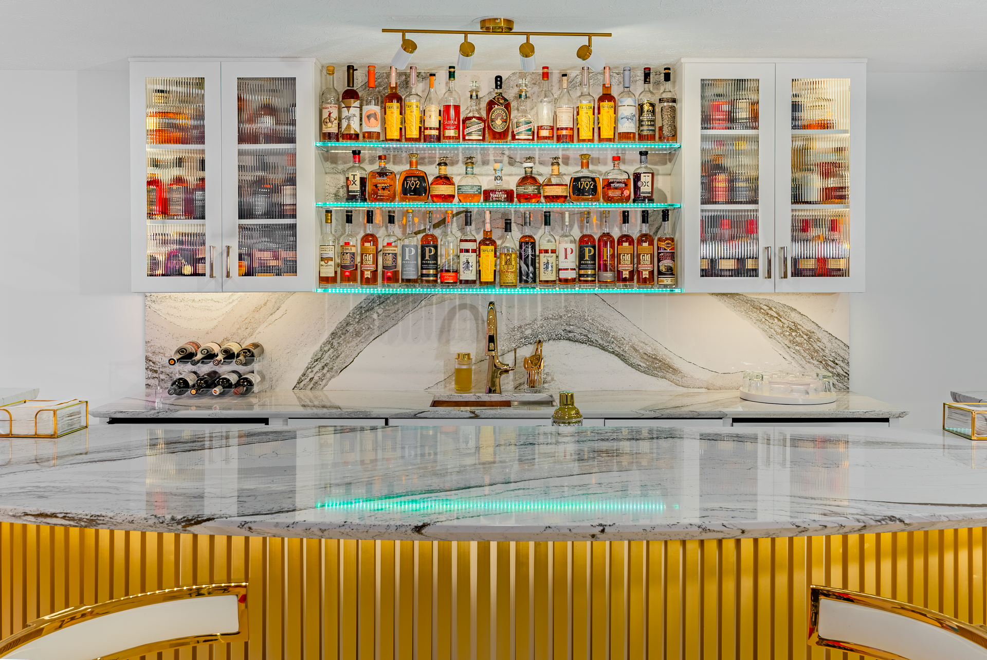 Bar with gold accents, stocked with liquor bottles. Marble countertop, mirrored backsplash, bright lighting.