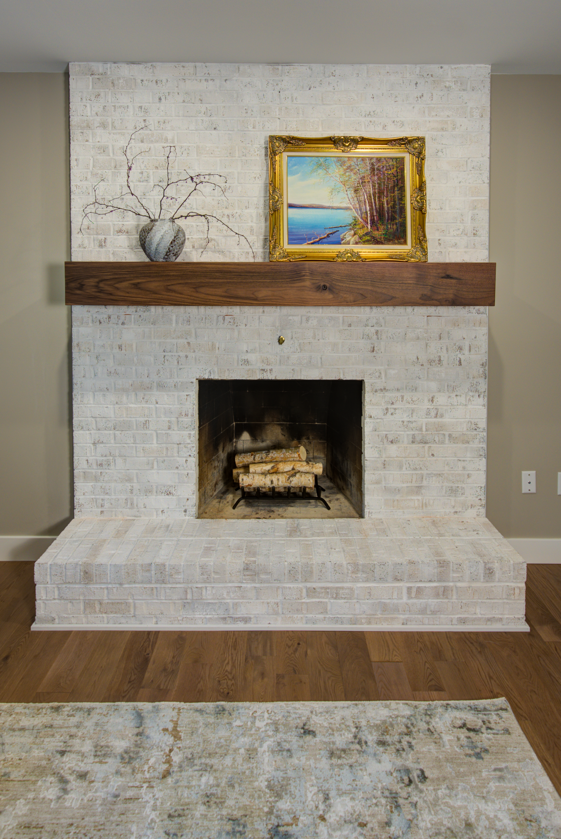 Fireplace with white brick, wooden mantel, artwork, and firewood.
