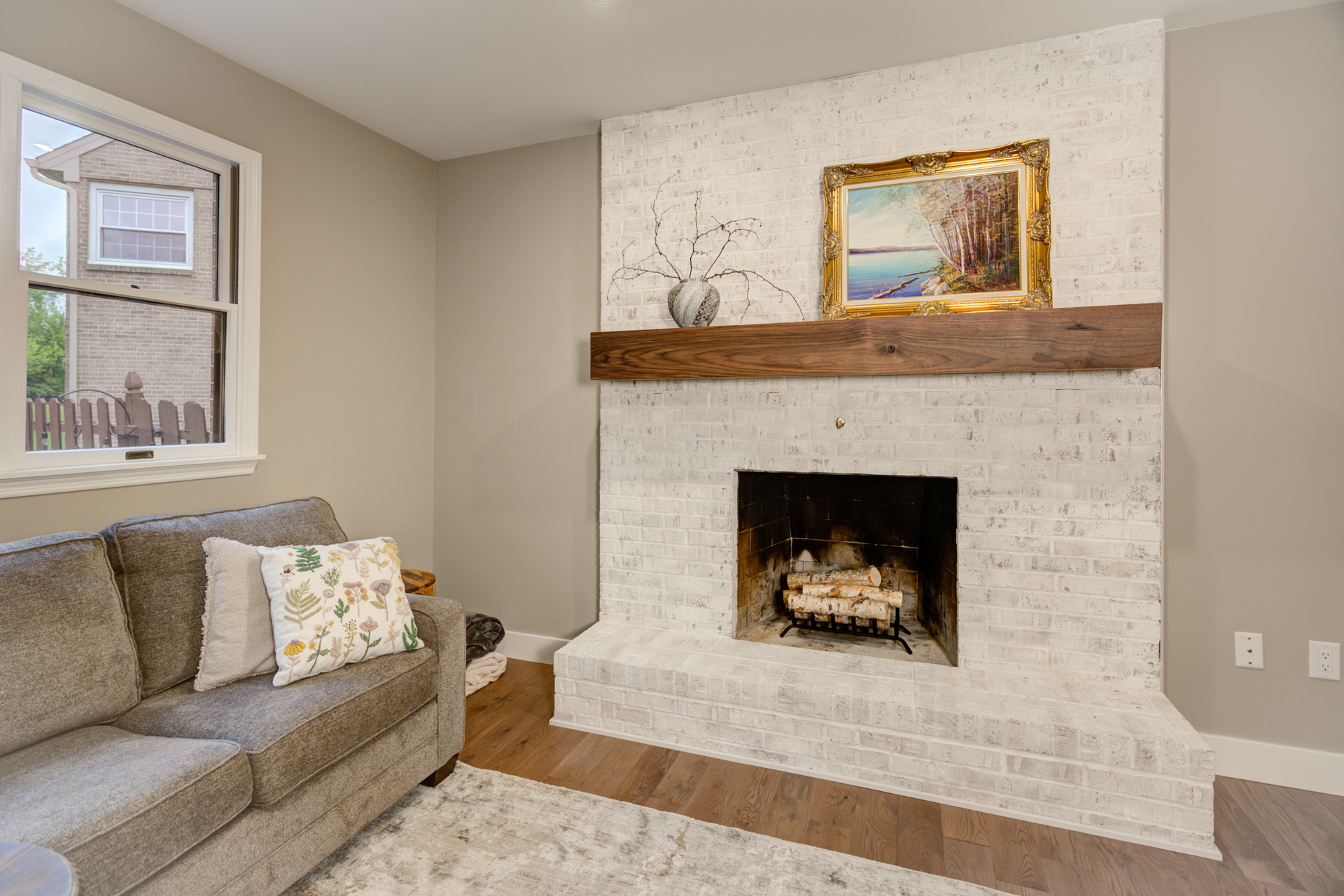 Living room with white brick fireplace, brown wooden mantel, artwork, and gray couch.