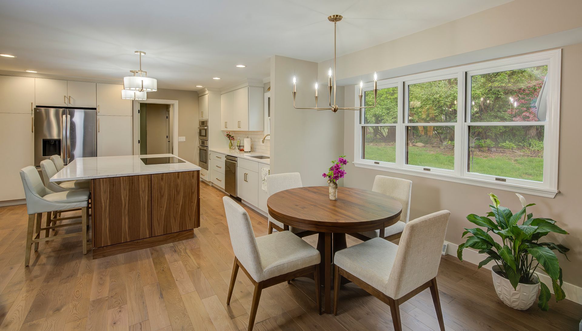 Modern kitchen with island, dining area, and large windows overlooking a backyard. Wooden floors, white cabinets, light fixtures.