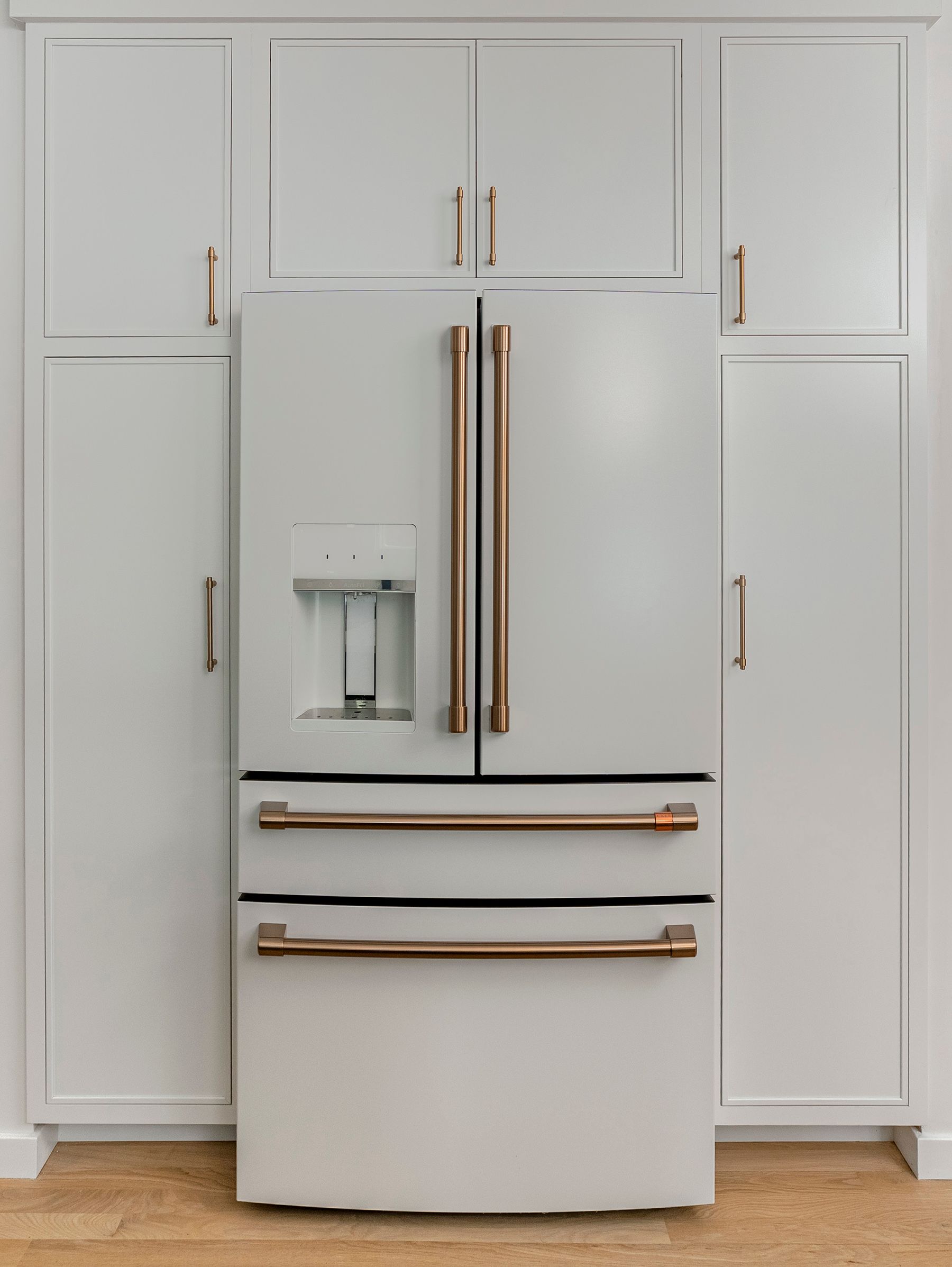 White refrigerator with bronze handles and trim, flanked by white cabinets, set in a kitchen.