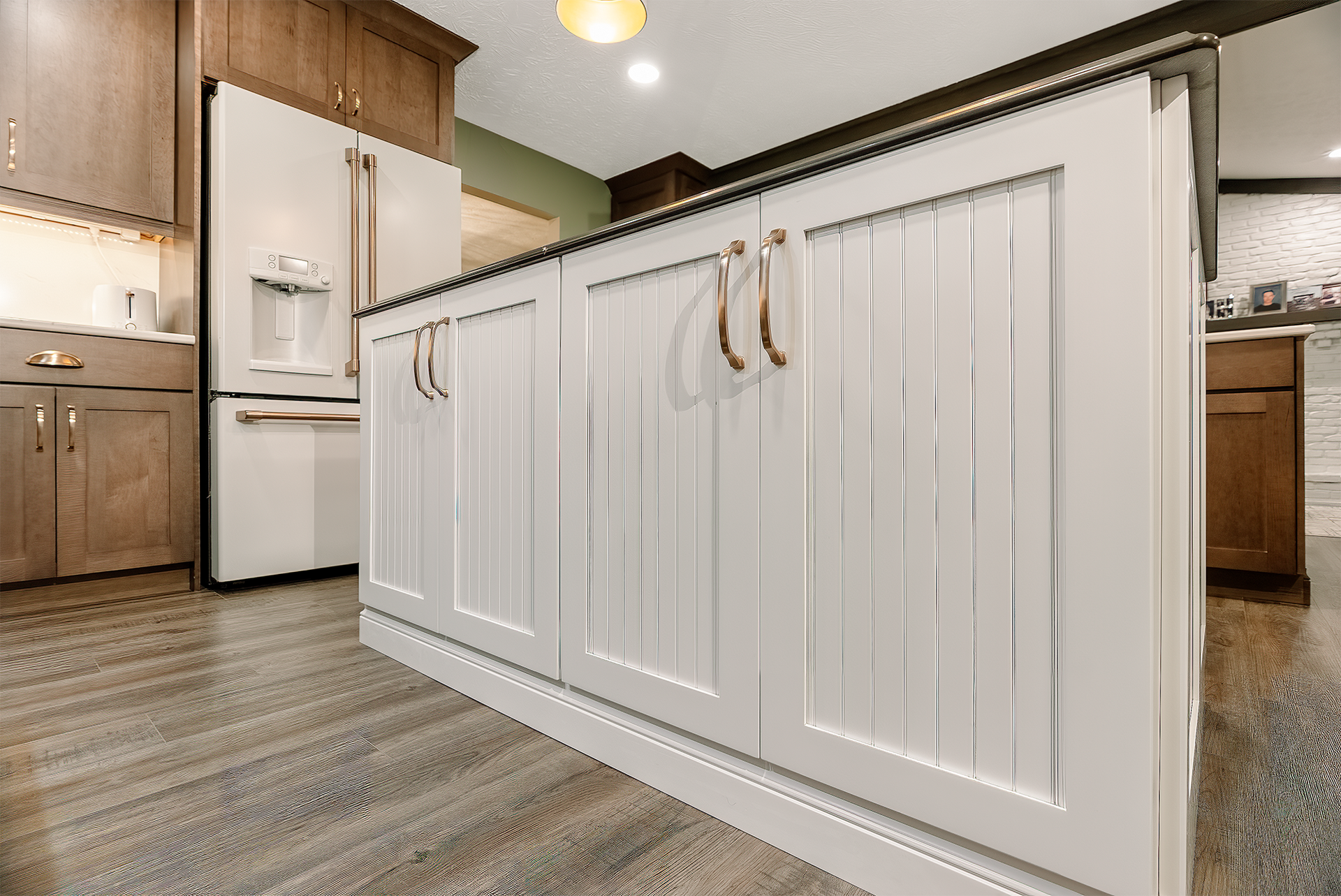 White kitchen island with copper handles and wood paneling. Gray flooring and white refrigerator.