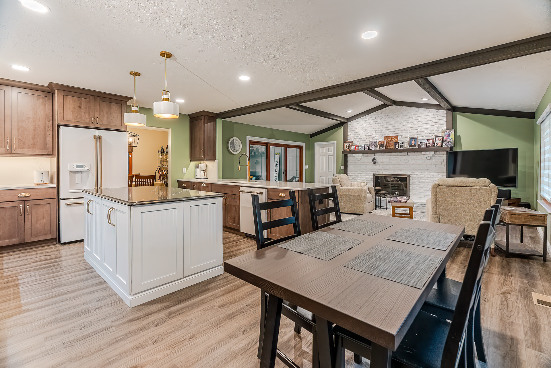 Open-concept kitchen and living room with brown cabinets, white island, dining table, fireplace, and large TV.