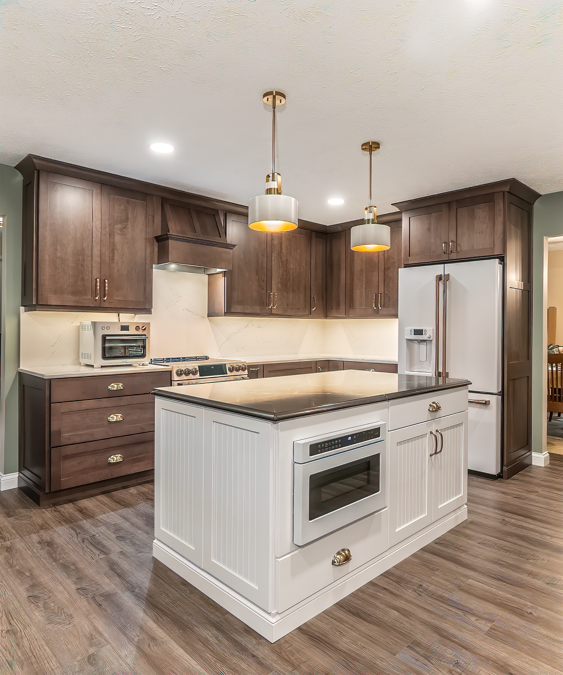 Kitchen with dark cabinets, white island, appliances. Gold pendant lights hang above the island.