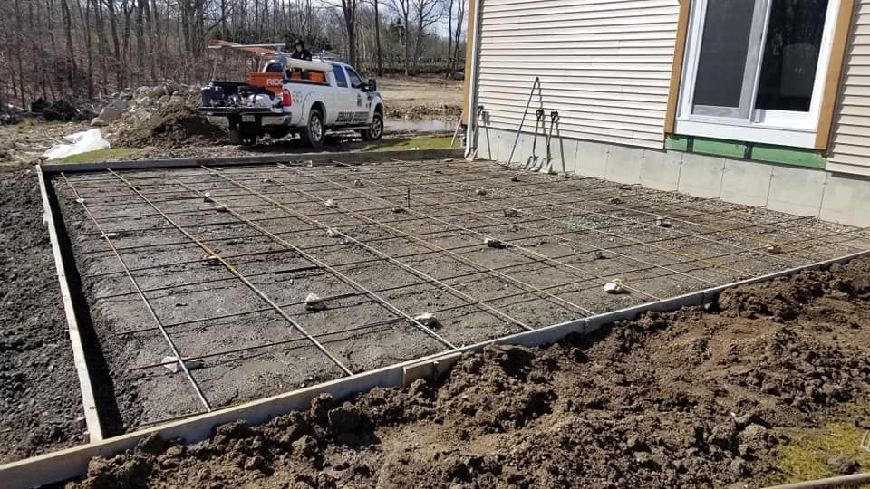Concrete patio under construction with rebar grid and wooden formwork next to a house and truck.