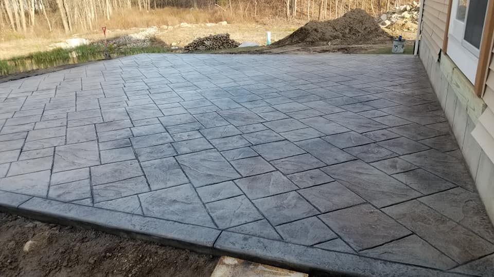 Stamped concrete patio with a border, next to a house, dirt in the foreground.